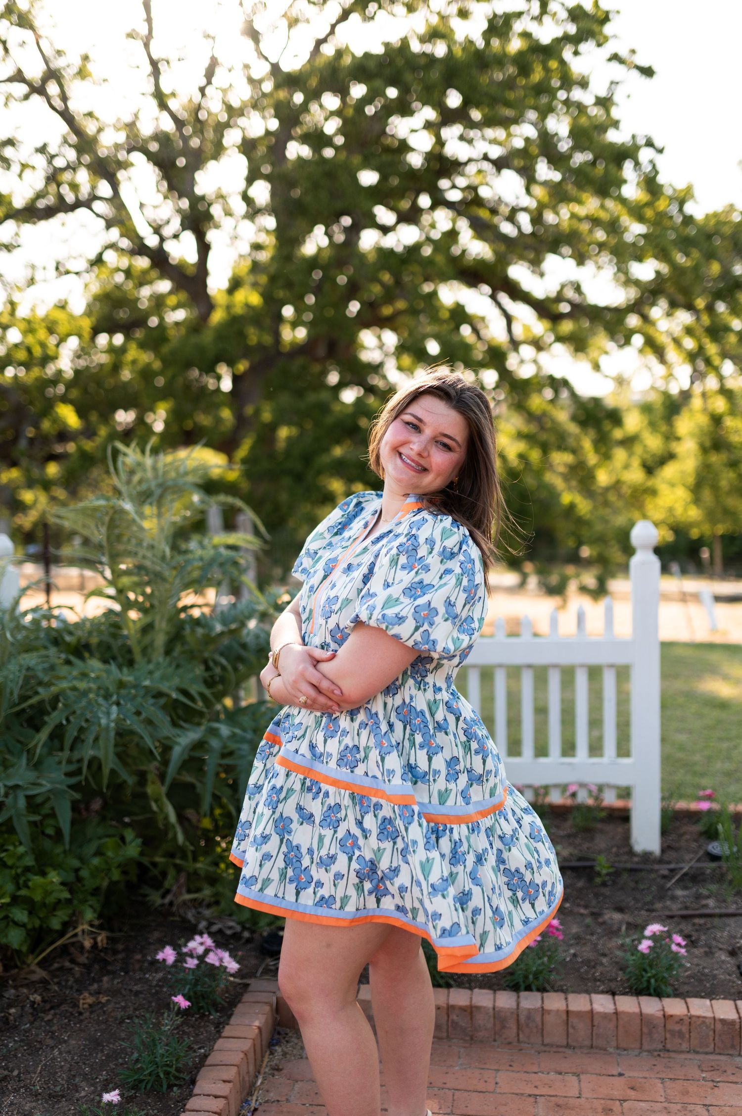 Happy woman in blue floral dress with orange trim twirling in garden near white fence.