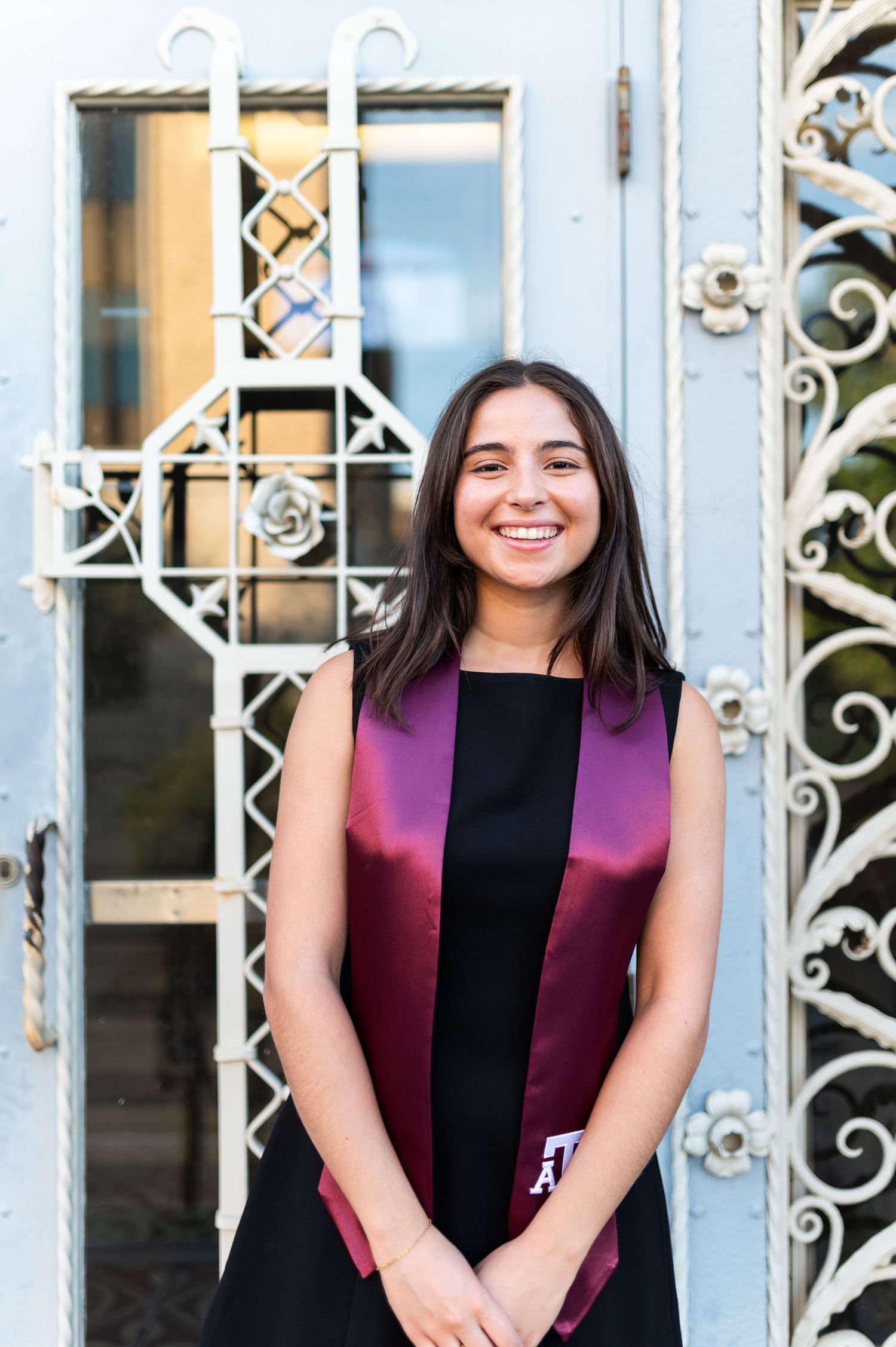 Smiling young woman in black dress with maroon Texas A&M graduation stole standing by ornate gate.