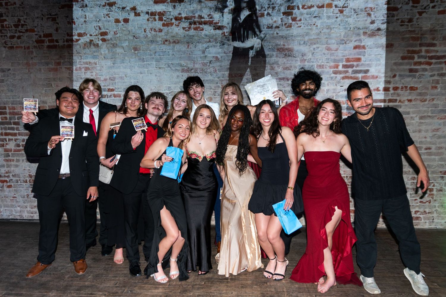 A group of elegantly dressed students pose together holding awards at a formal event against a brick wall backdrop.