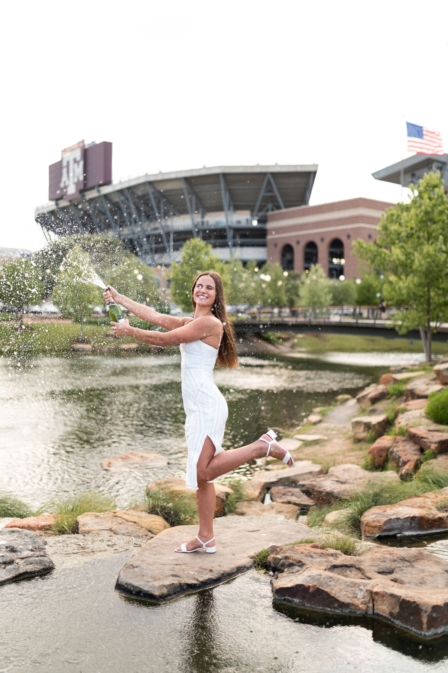 Smiling Texas A&M female graduate pops champagne on rocks near campus pond with Kyle Field in background.