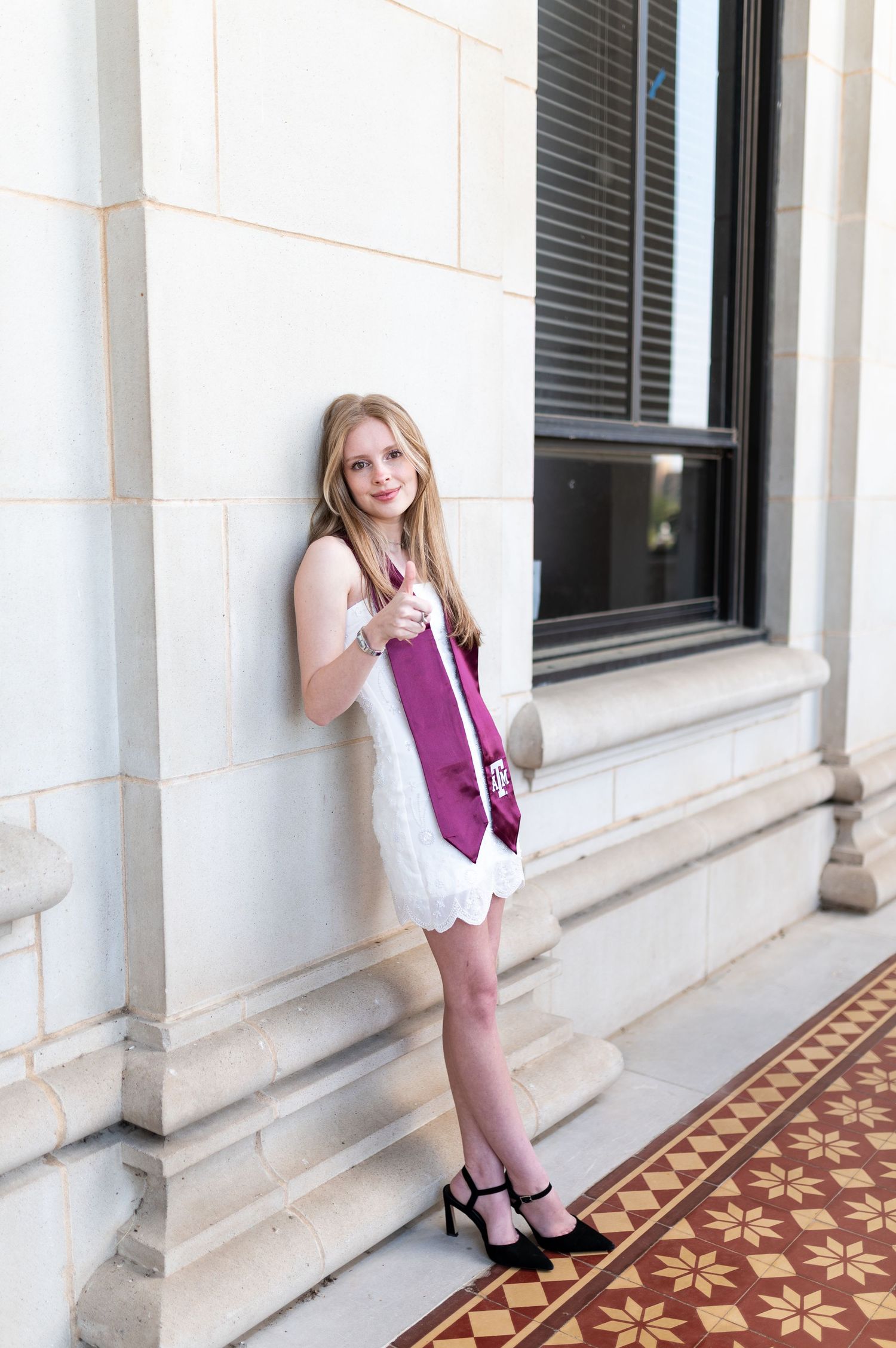 Blonde graduate in white dress with Texas A&M maroon stole leans against stone column giving thumbs up. Grad Photos at the Admin building