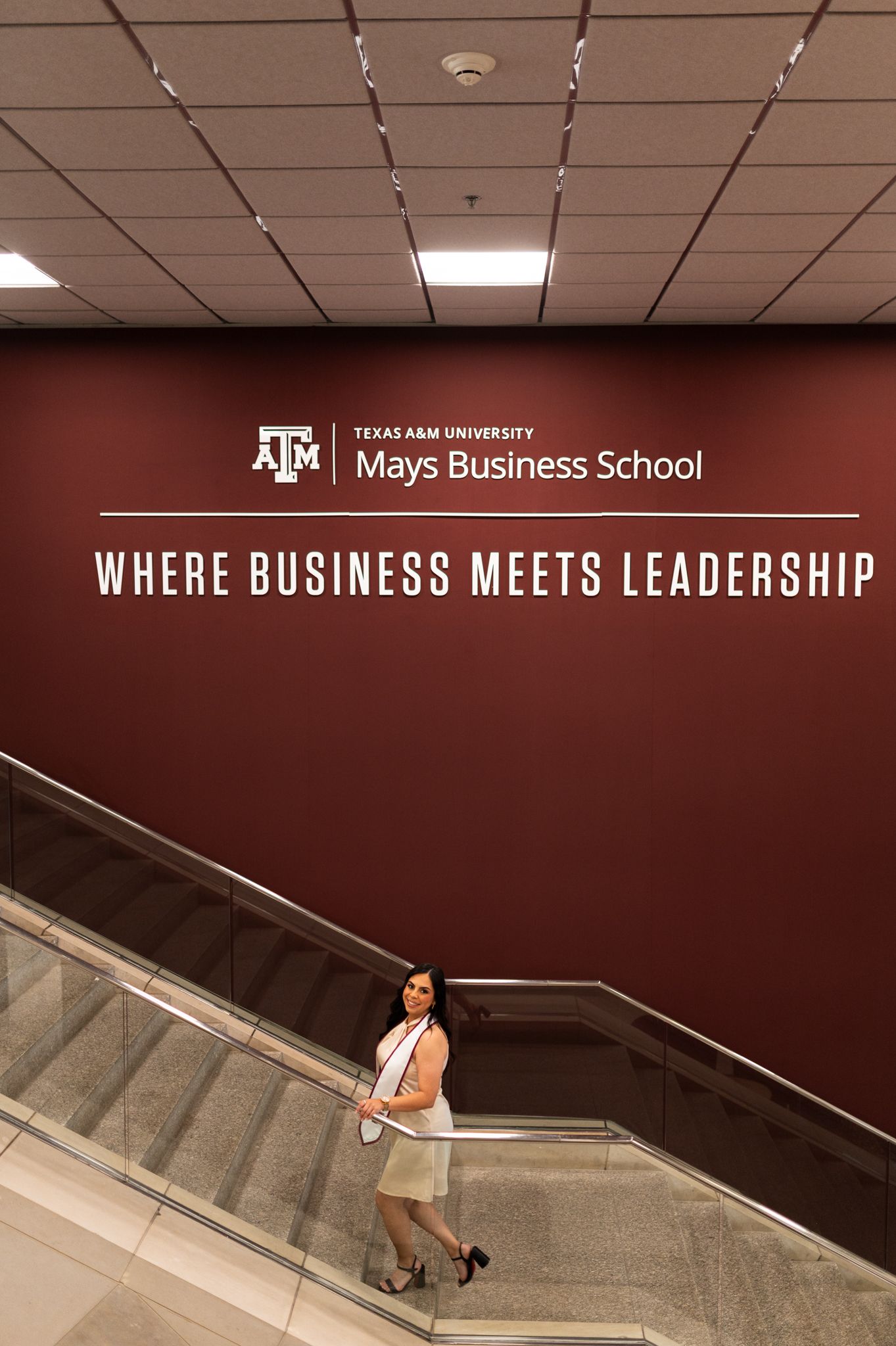 Texas A&M Mays Business School graduate stands on escalator beneath maroon Where Business Meets Leadership sign.