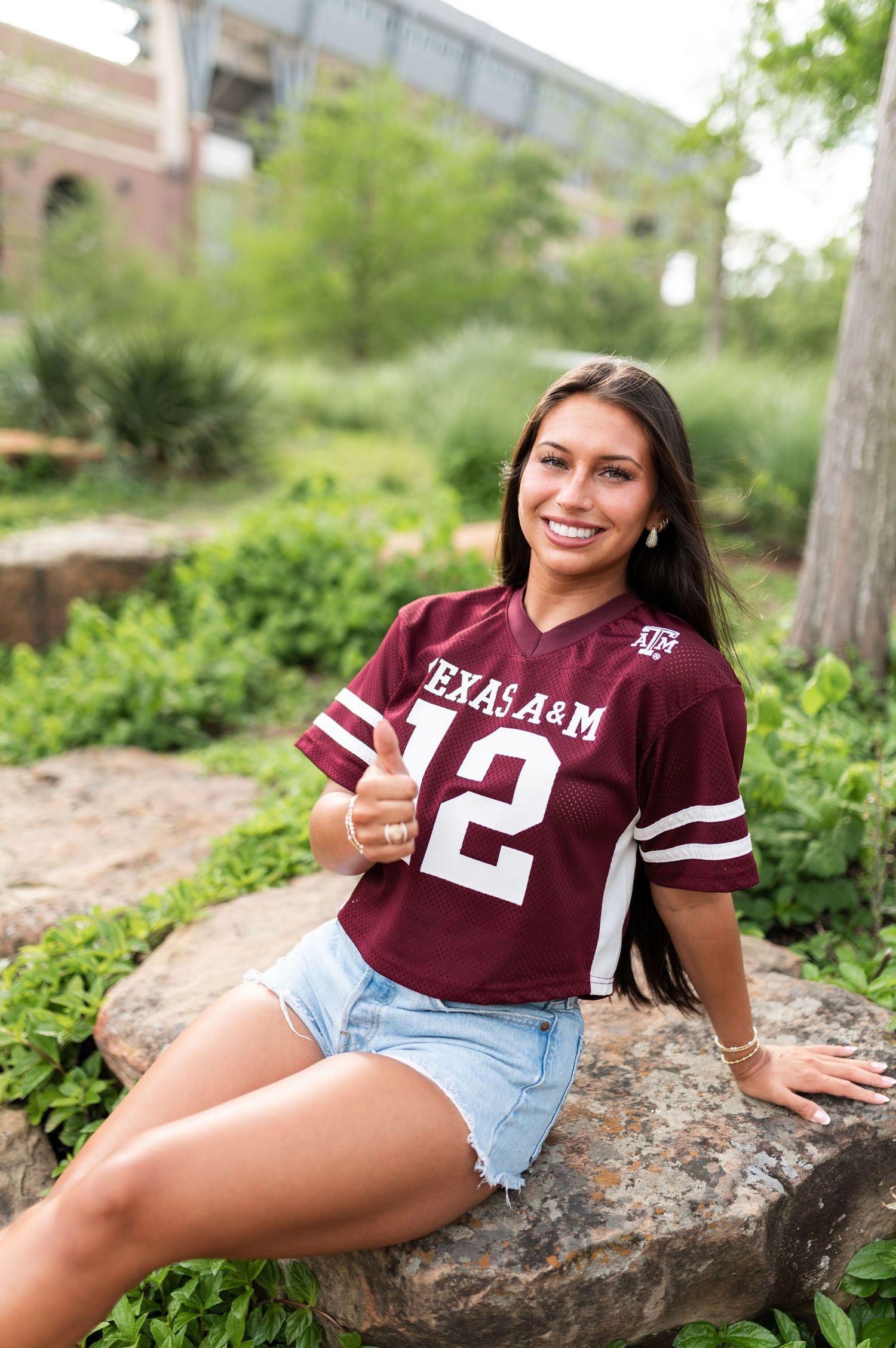 A smiling young woman in a Texas A&M jersey giving a thumbs up while seated on rocks.
