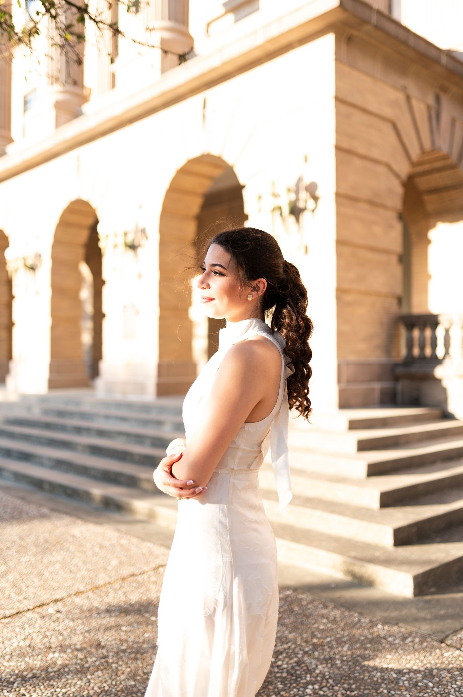 Woman in white halter dress poses gracefully outside sunlit stone building.