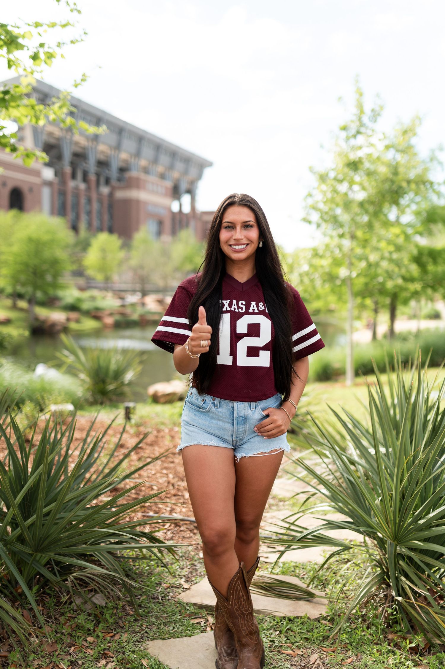 A smiling young woman in a Texas A&M jersey giving a thumbs up on a campus path.