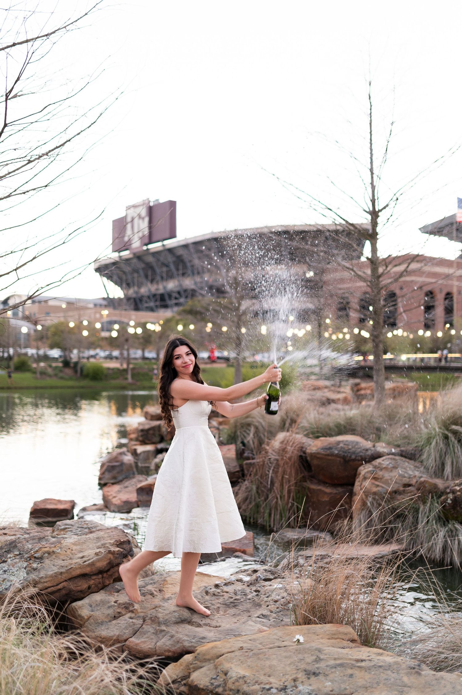 Woman in white dress joyfully pops champagne bottle outdoors near Texas A&M stadium at dusk.