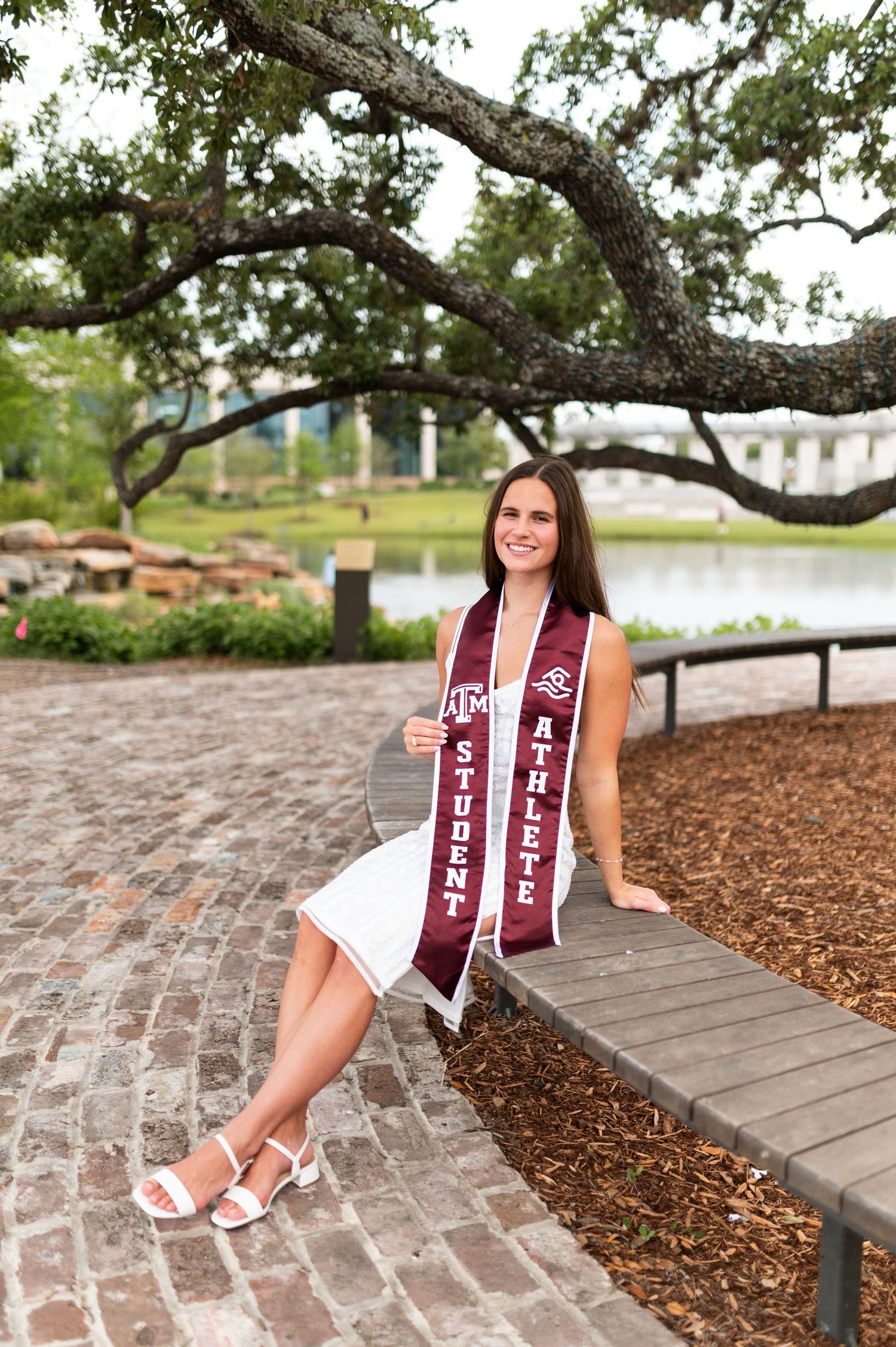 Texas A&M female student athlete in white dress displays Student Athlete sash outdoors near campus pond.