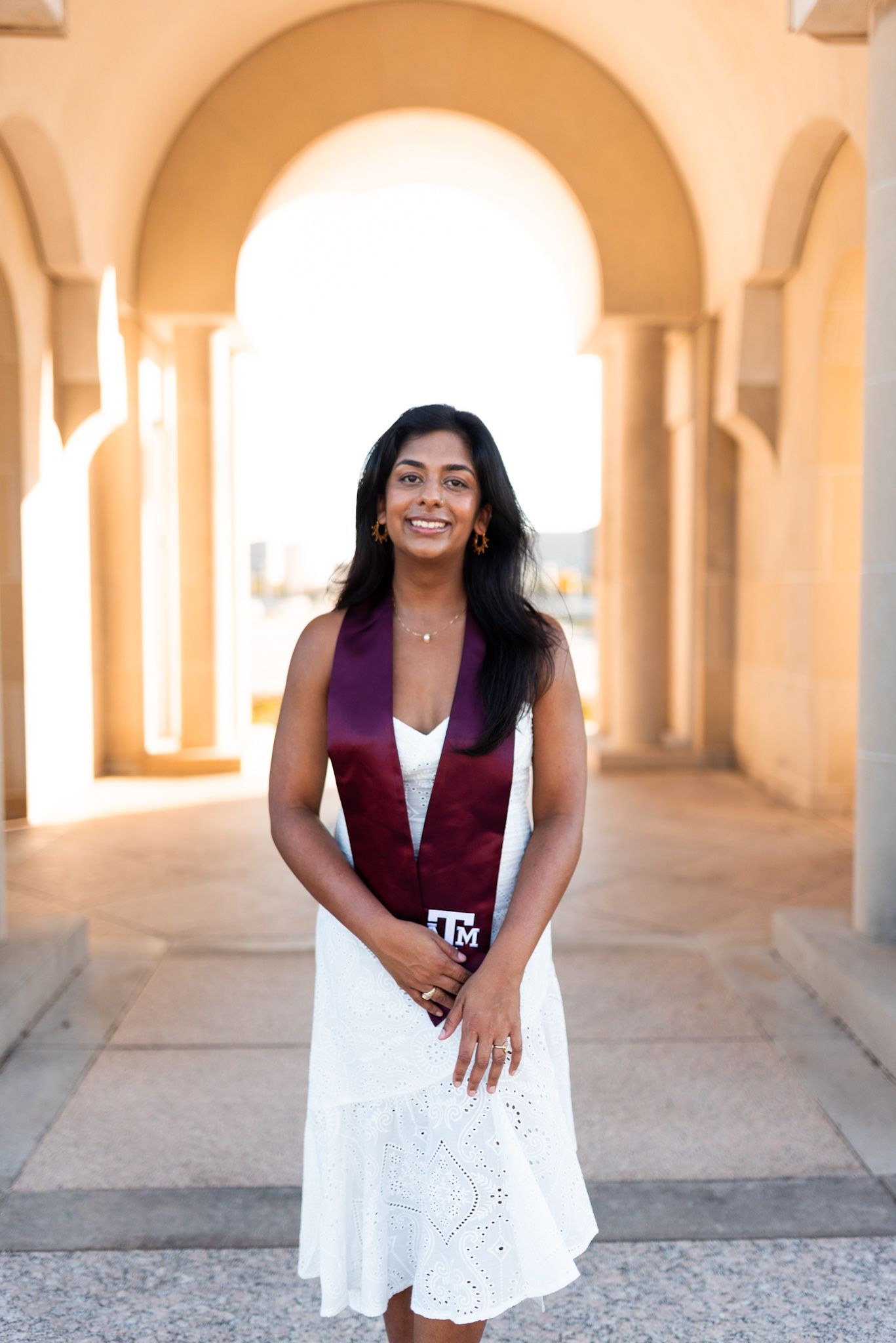 Graduate in white dress wearing Texas A&M maroon stole standing under stone arches.