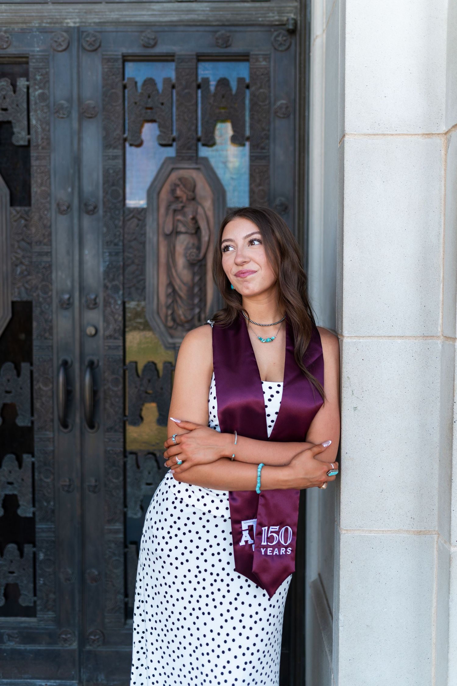 Female Texas A&M graduate leans against stone column wearing maroon 150 Years stole and white polka dot dress. Grad Photos at the Admin