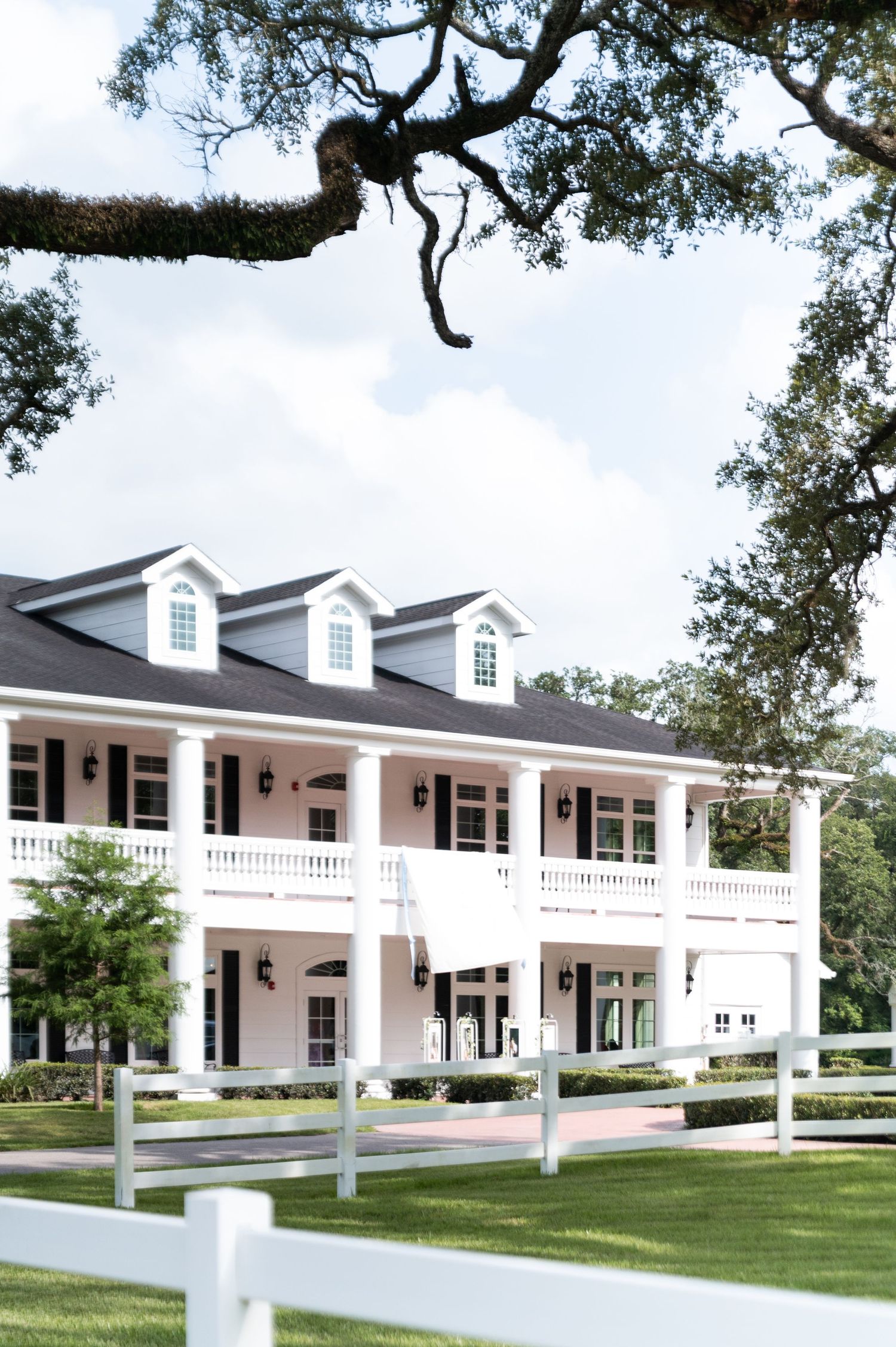Elegant white Southern plantation-style wedding venue surrounded by oak trees and white fence on a sunny day.