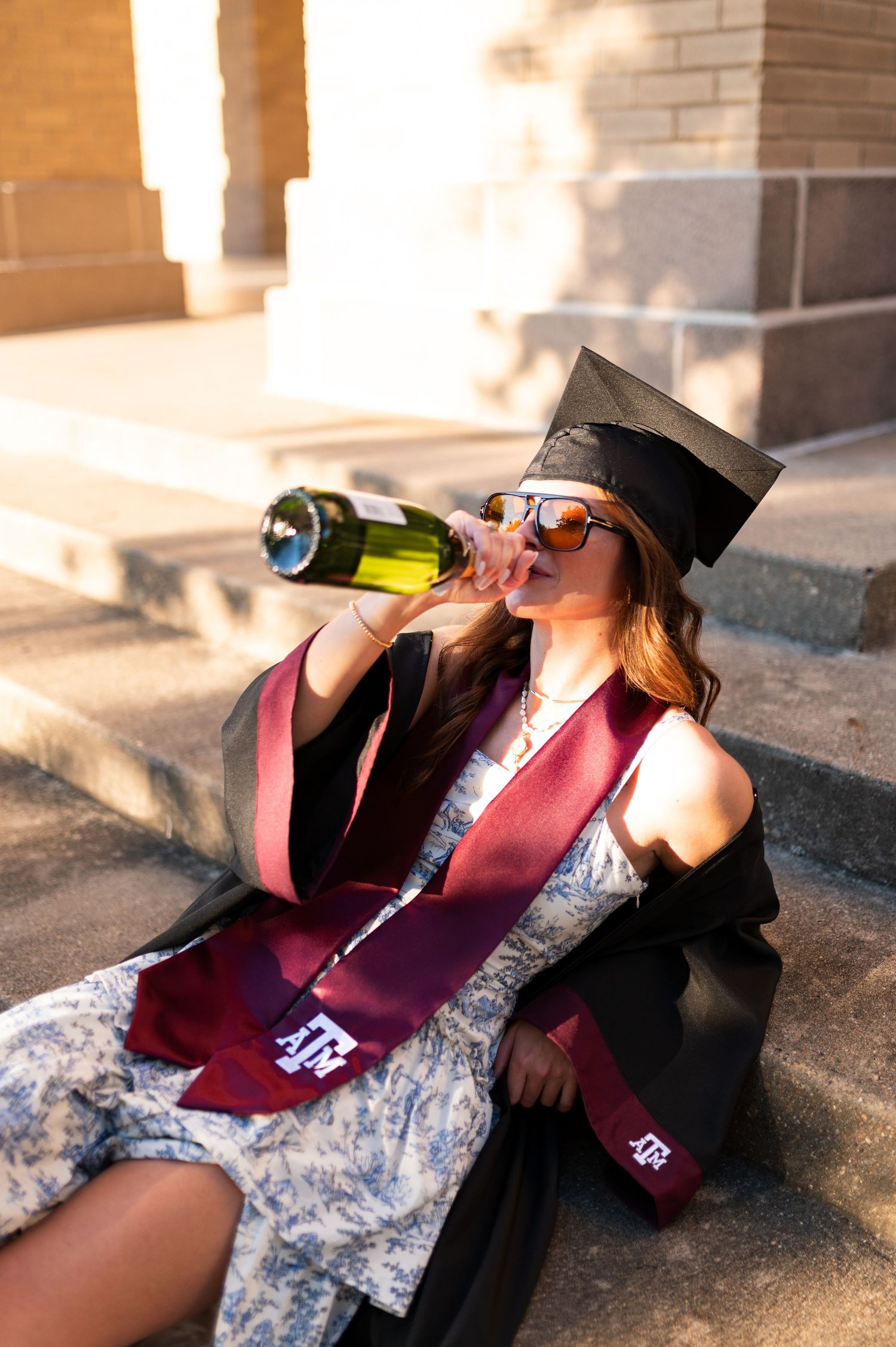 Texas A&M graduate in cap and gown drinks from wine bottle on steps wearing sunglasses and maroon stole.