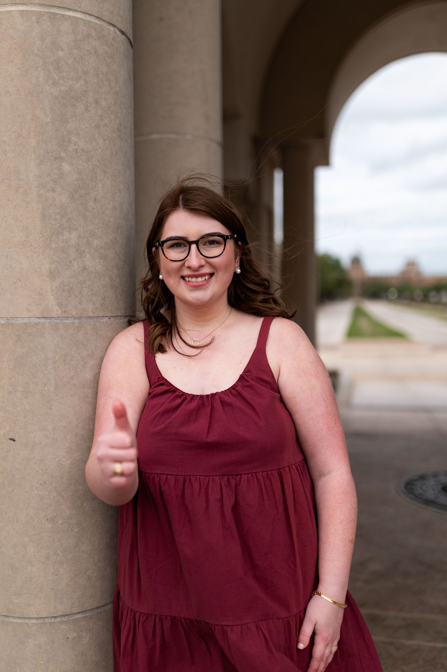 Smiling Texas A&M graduate in maroon dress giving thumbs up while leaning on stone column.