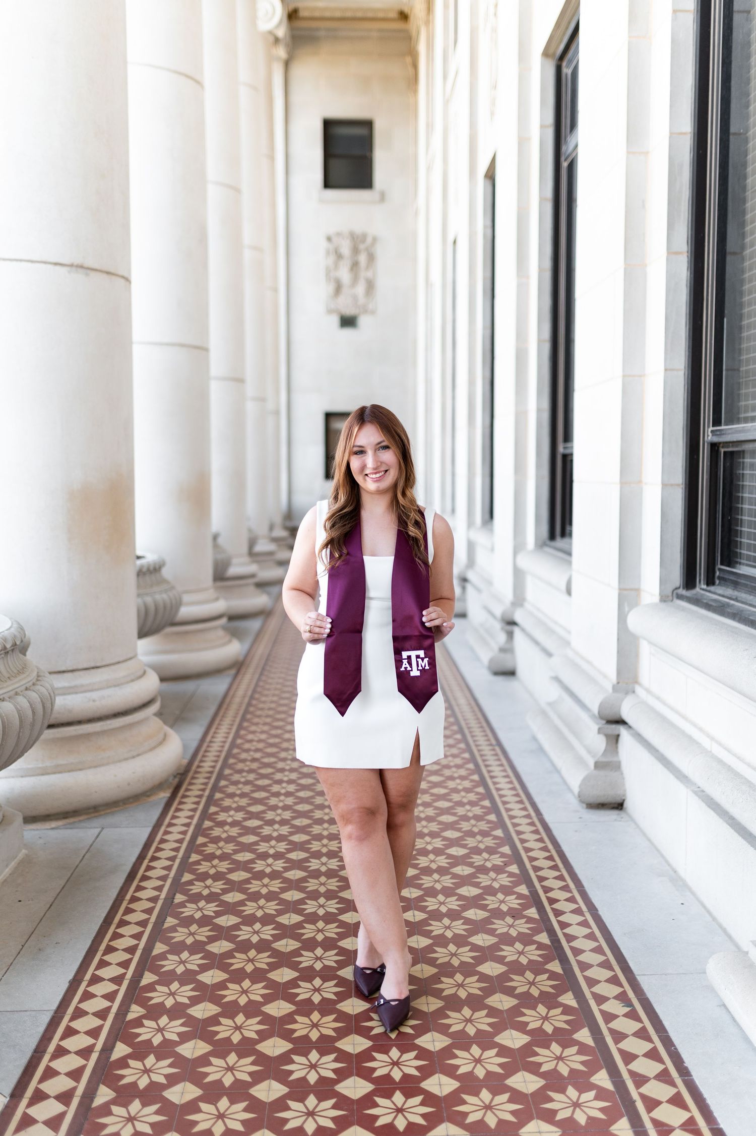 Graduate in white dress wearing Texas A&M maroon stole walking through elegant columned hallway. Texas A&M Grad Photos at the Admin building