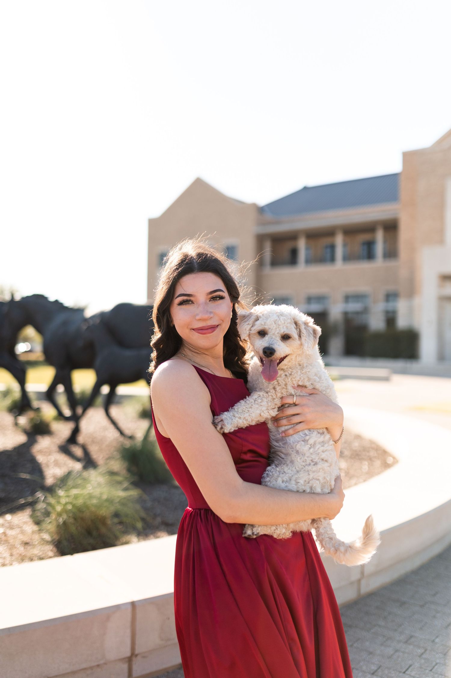 Smiling young woman in red dress holding a fluffy white dog outdoors near horse sculptures.