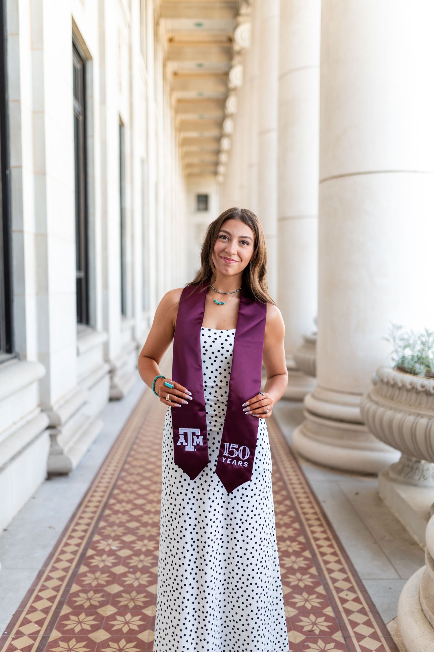 Smiling woman holds Texas A&M 150 Years maroon graduation stole in colonnade. Grad Photos at the Admin building