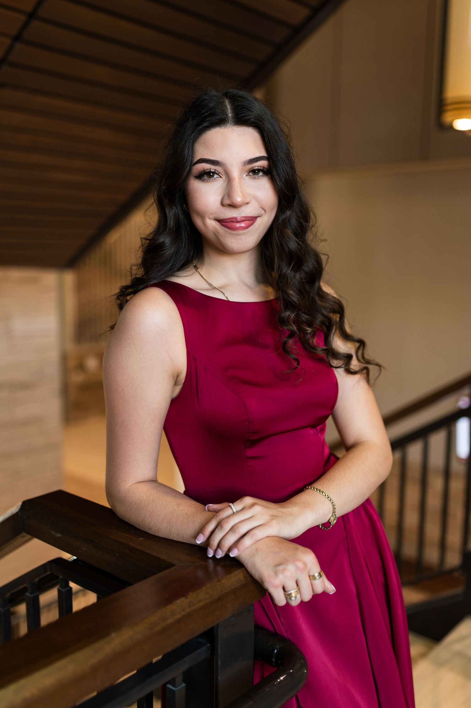 Young woman in elegant maroon dress leaning on wooden railing in a warm indoor setting.