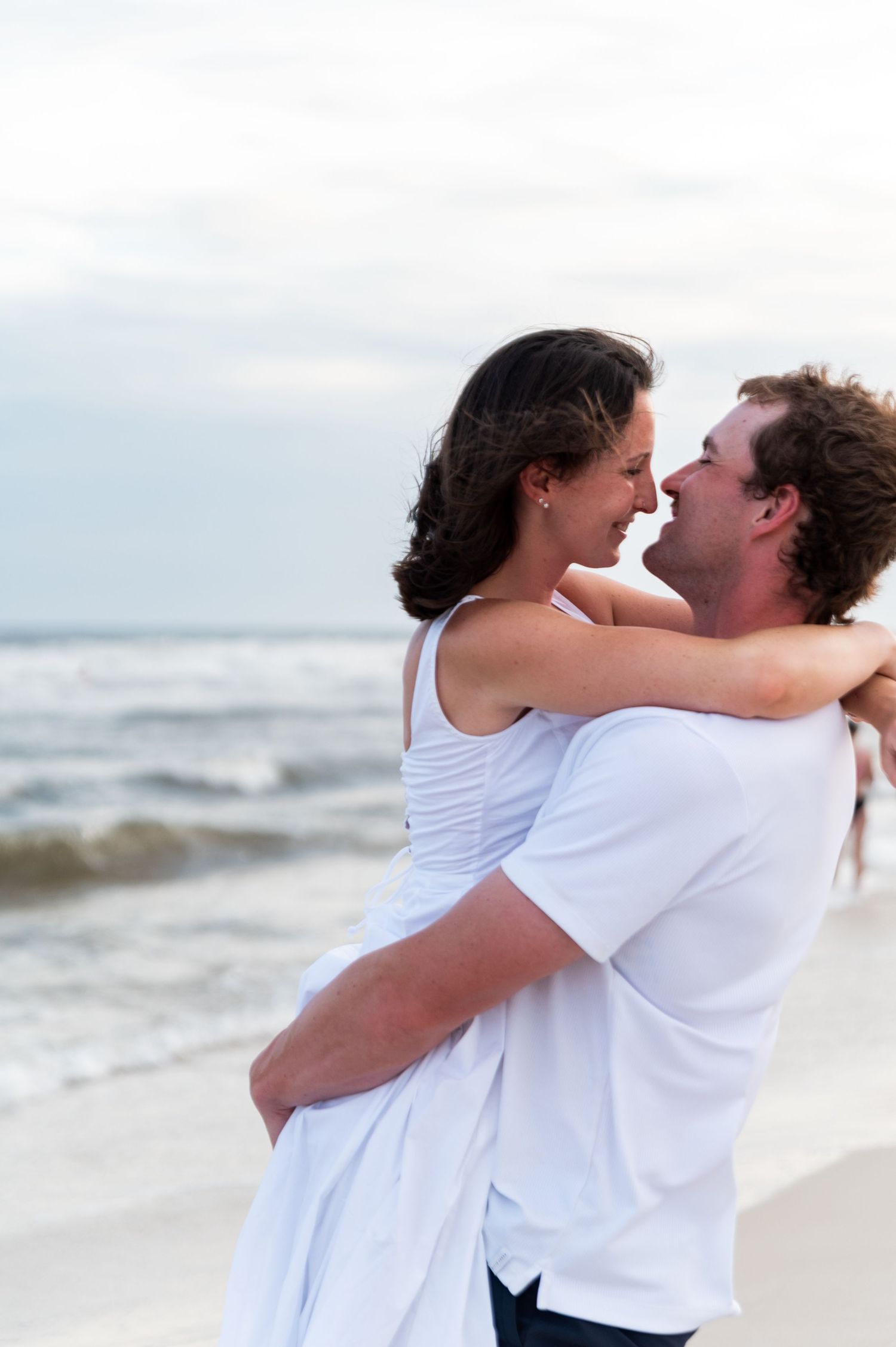 A happy couple embraces and smiles at each other on a beach with ocean waves in the background.