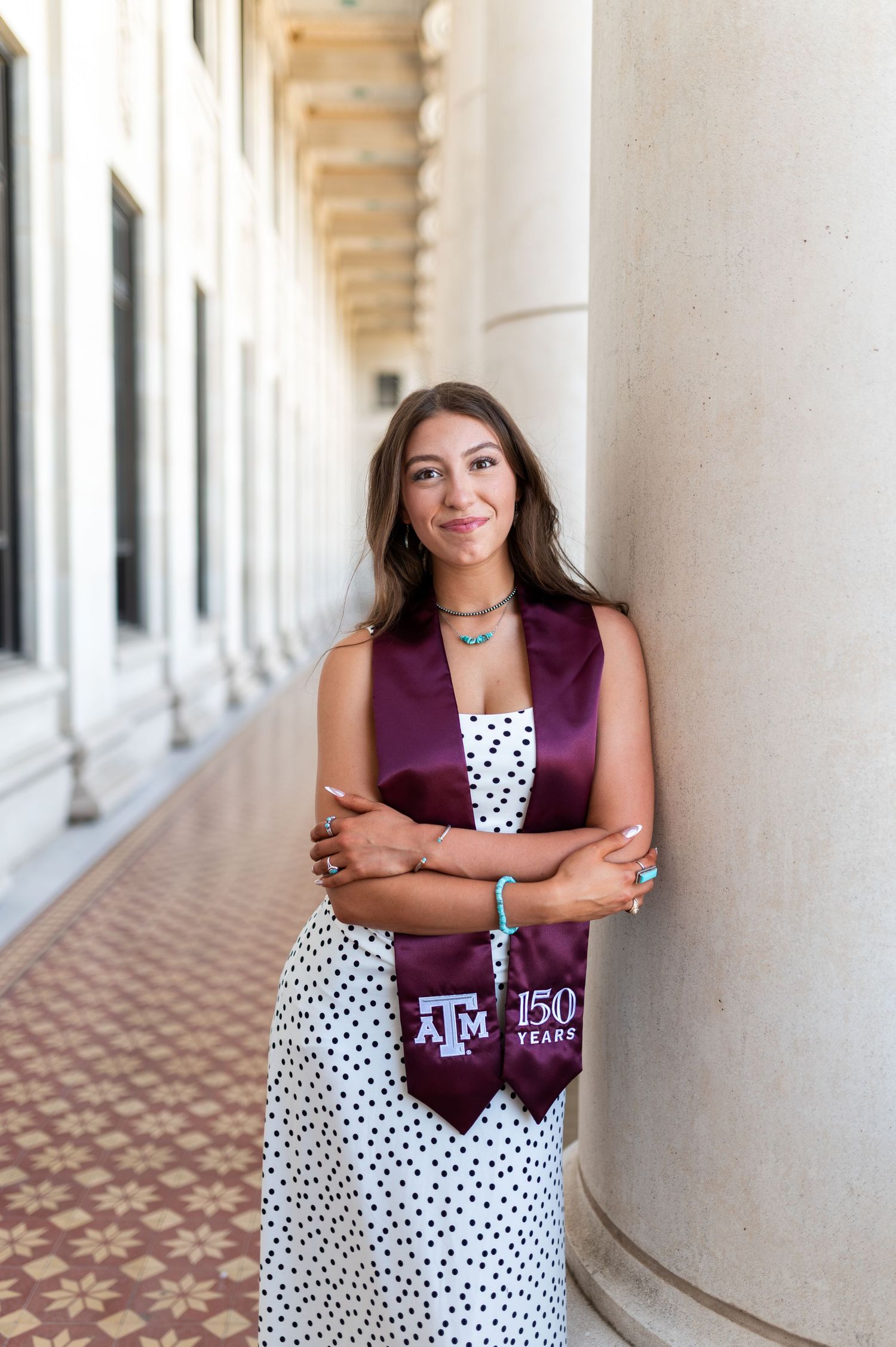 Woman in polka dot dress with Texas A&M 150 Years stole leans on column. Grad Photos at the Admin building