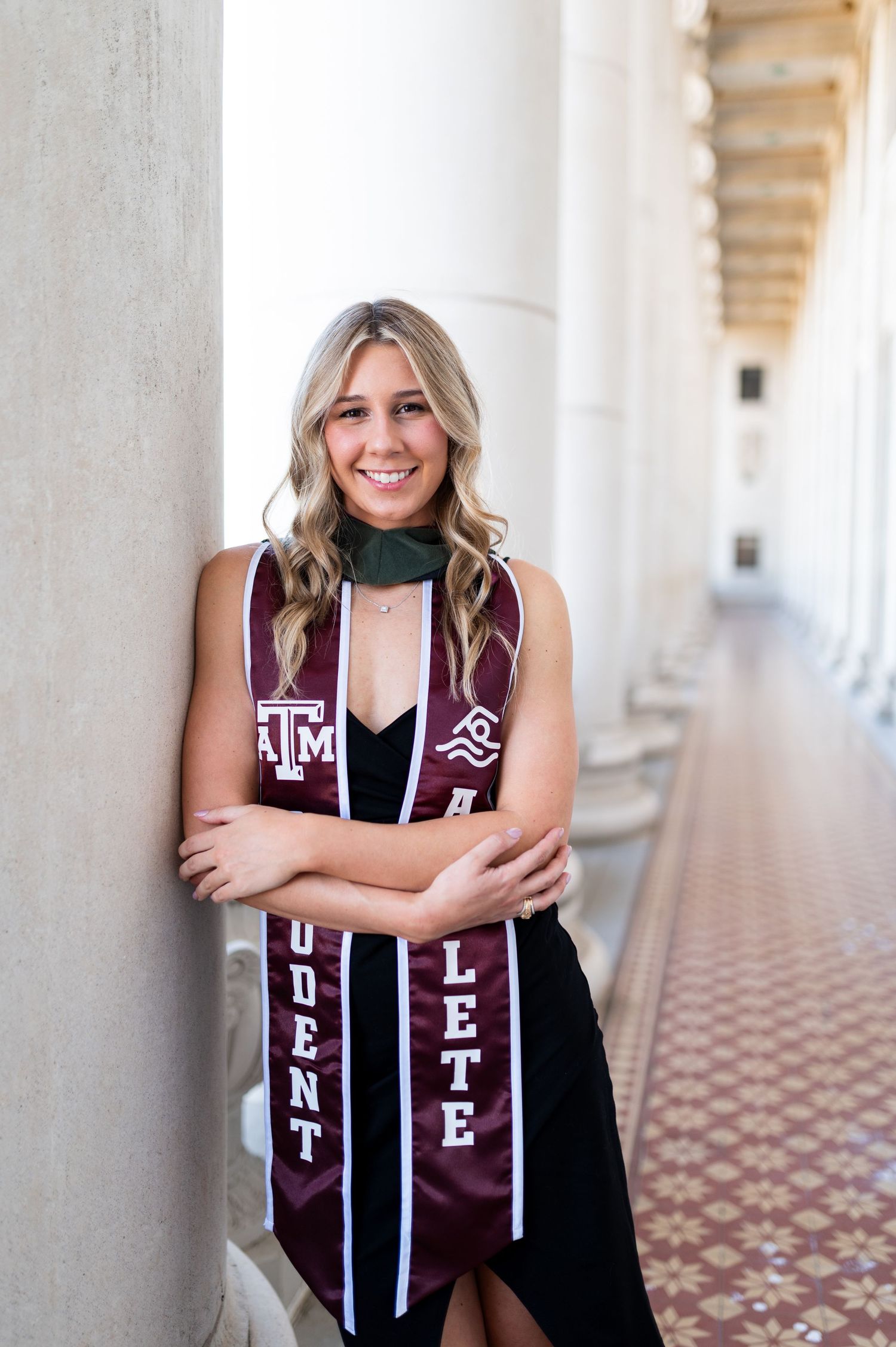 Smiling blonde woman wearing Texas A&M maroon student-athlete graduation stole leaning against column. Grad Photos at the Admin building