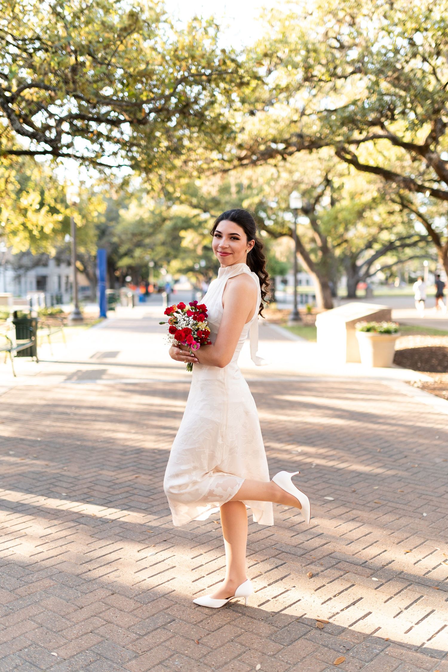 Joyful bride in white dress twirls with red bouquet on brick walkway beneath sunlit oak trees.