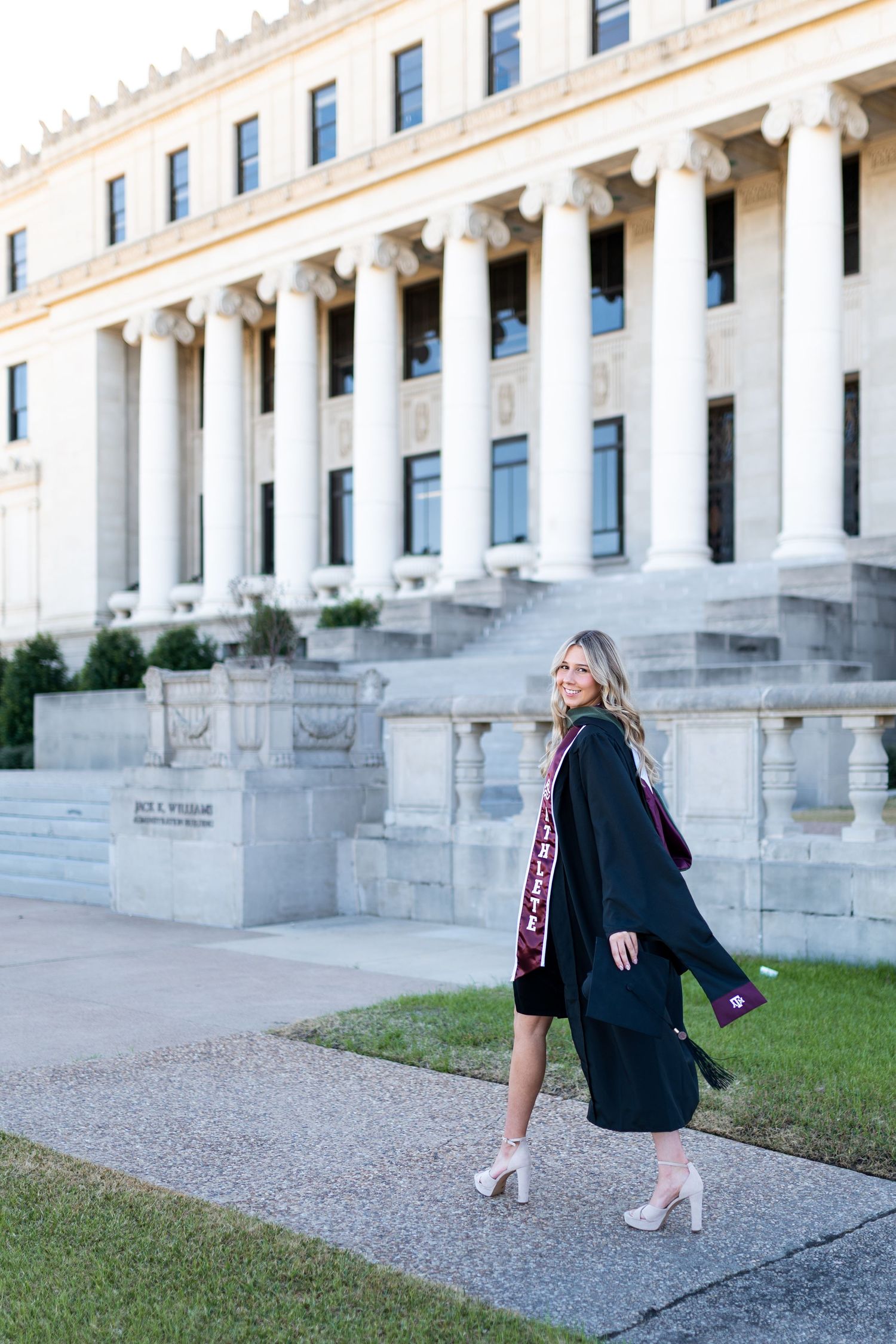 Texas A&M graduate walks in black gown past a grand white columned building on campus. Grad Photos at the Admin building