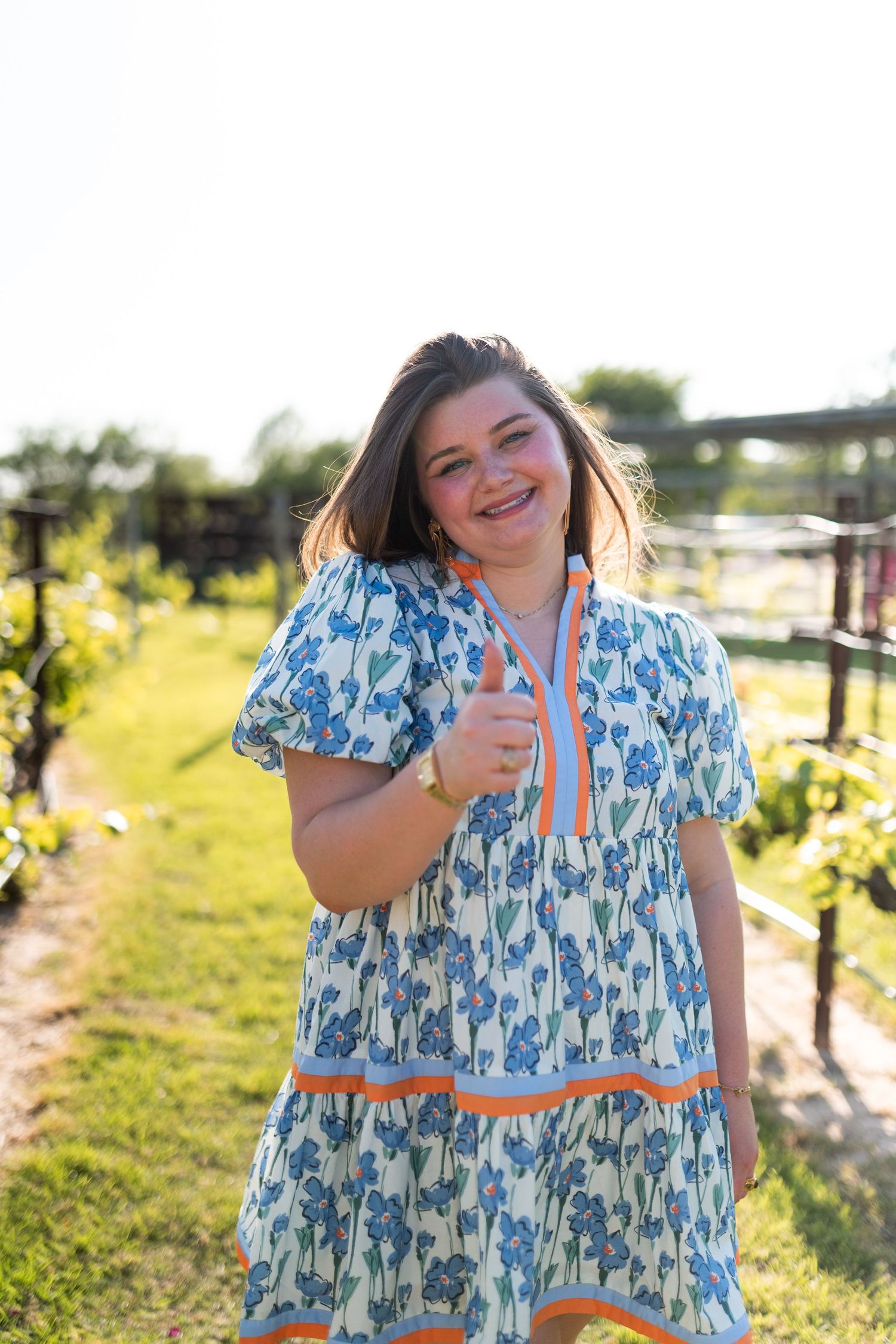 Cheerful woman in blue floral dress giving thumbs up in sunny garden vineyard setting.
