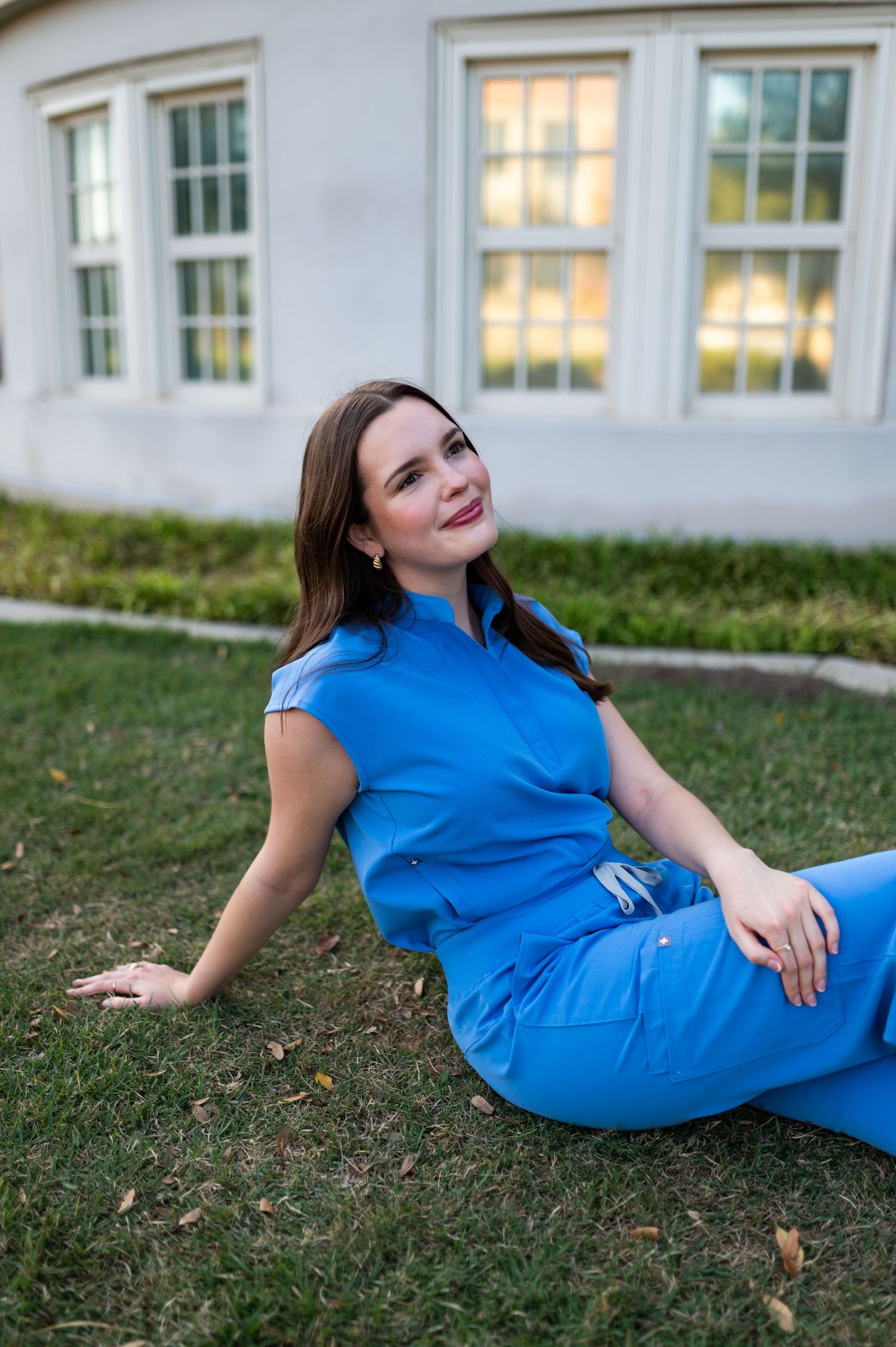 Young woman in blue outfit sits relaxed on green grass looking up near white building exterior.
