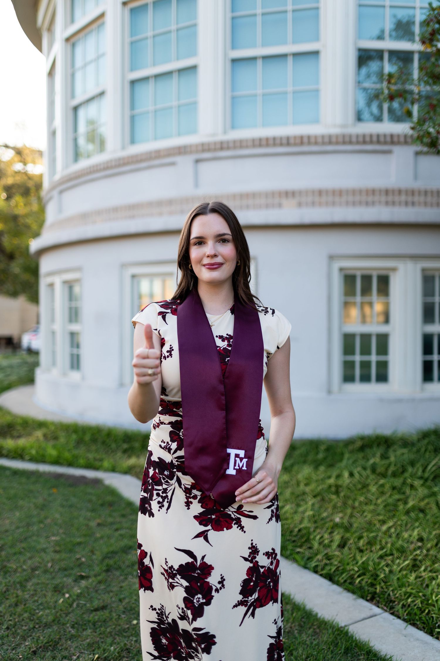 Texas A&M female graduate in floral dress gives thumbs up wearing maroon graduation stole outdoors.