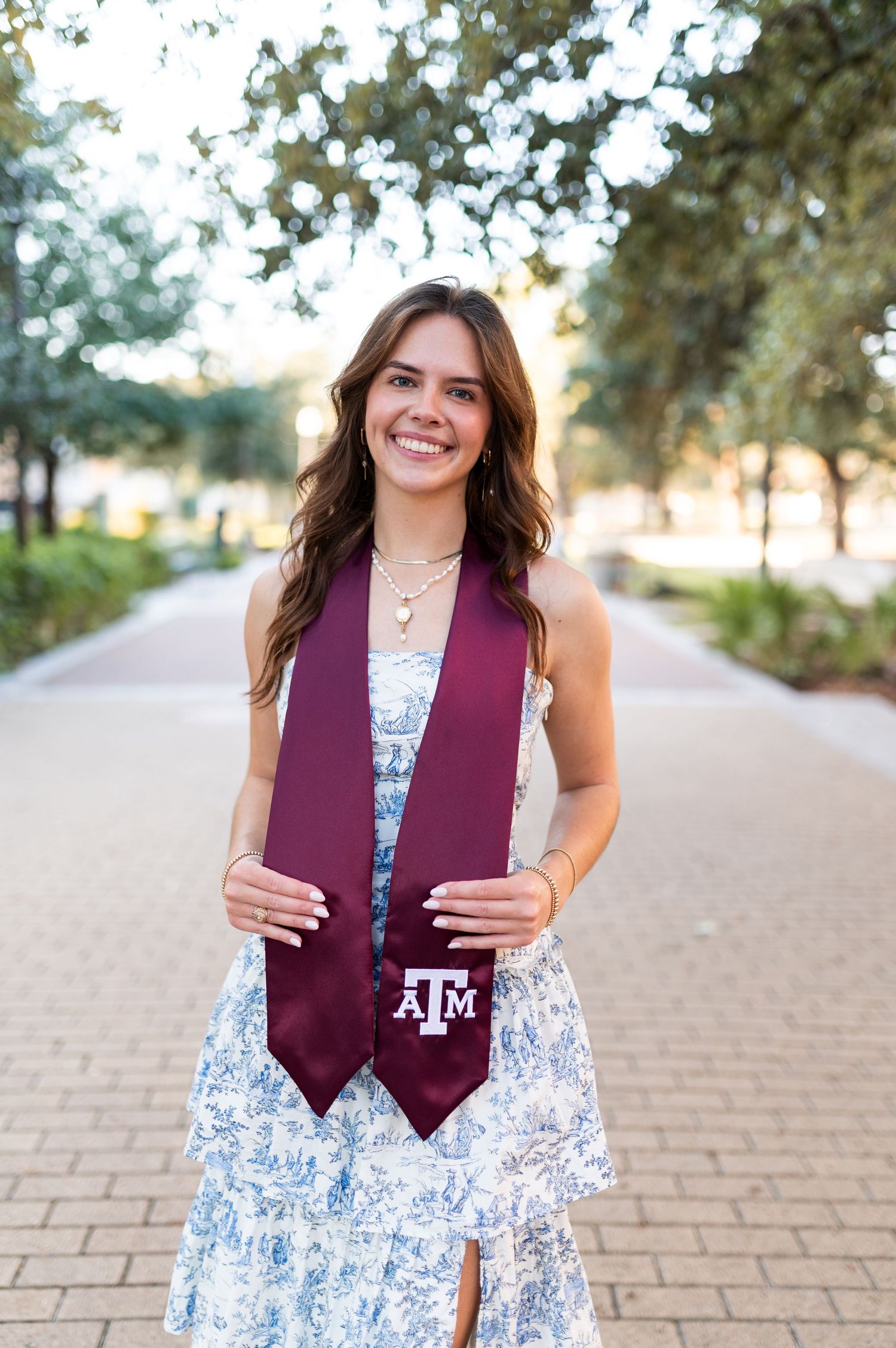 A happy young woman proudly displays her Texas A&M maroon graduation stole while standing on a sunlit campus walkway.