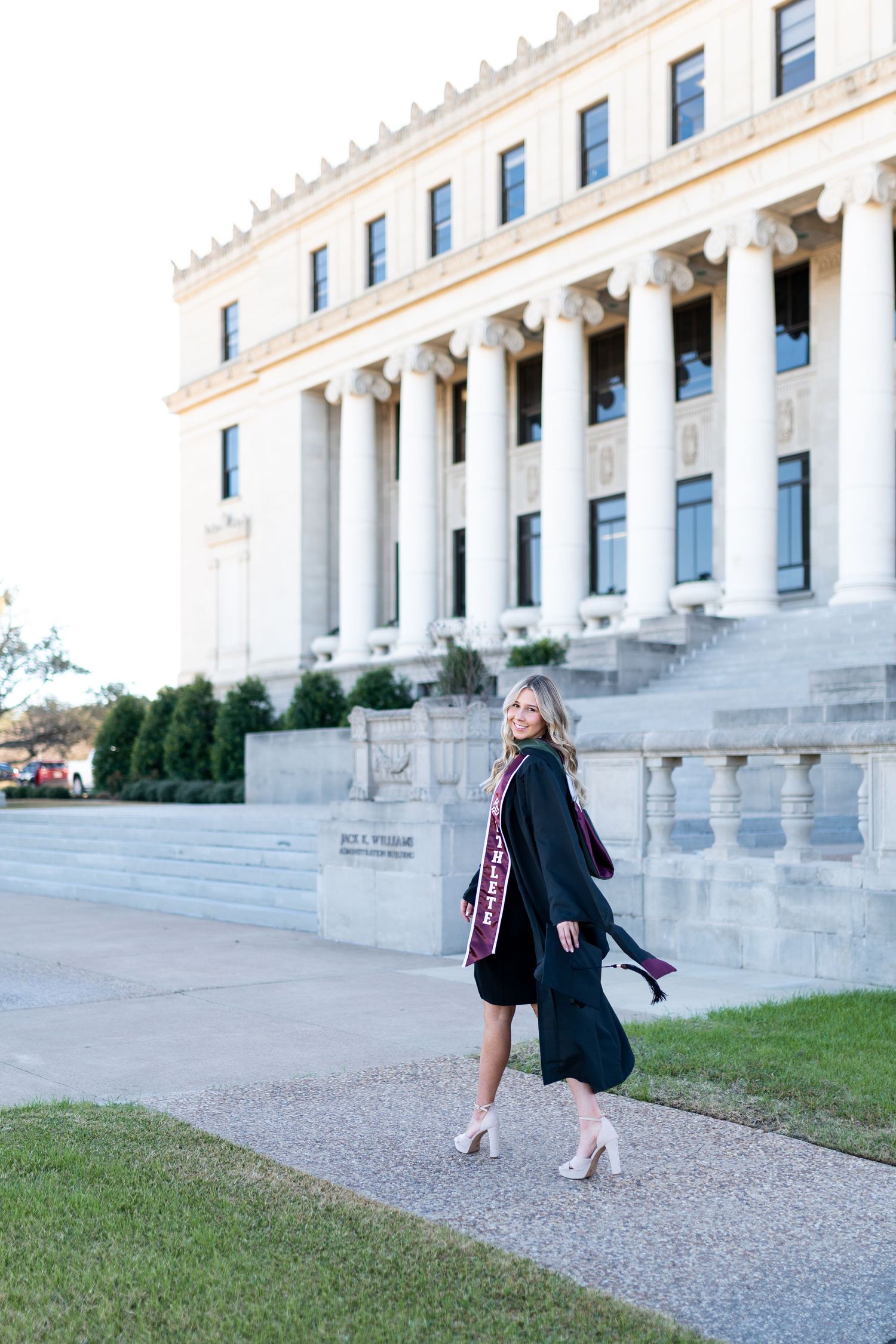 Woman in graduation gown walks outside a classic columned building at Texas A&M. Grad Photos at the Admin building