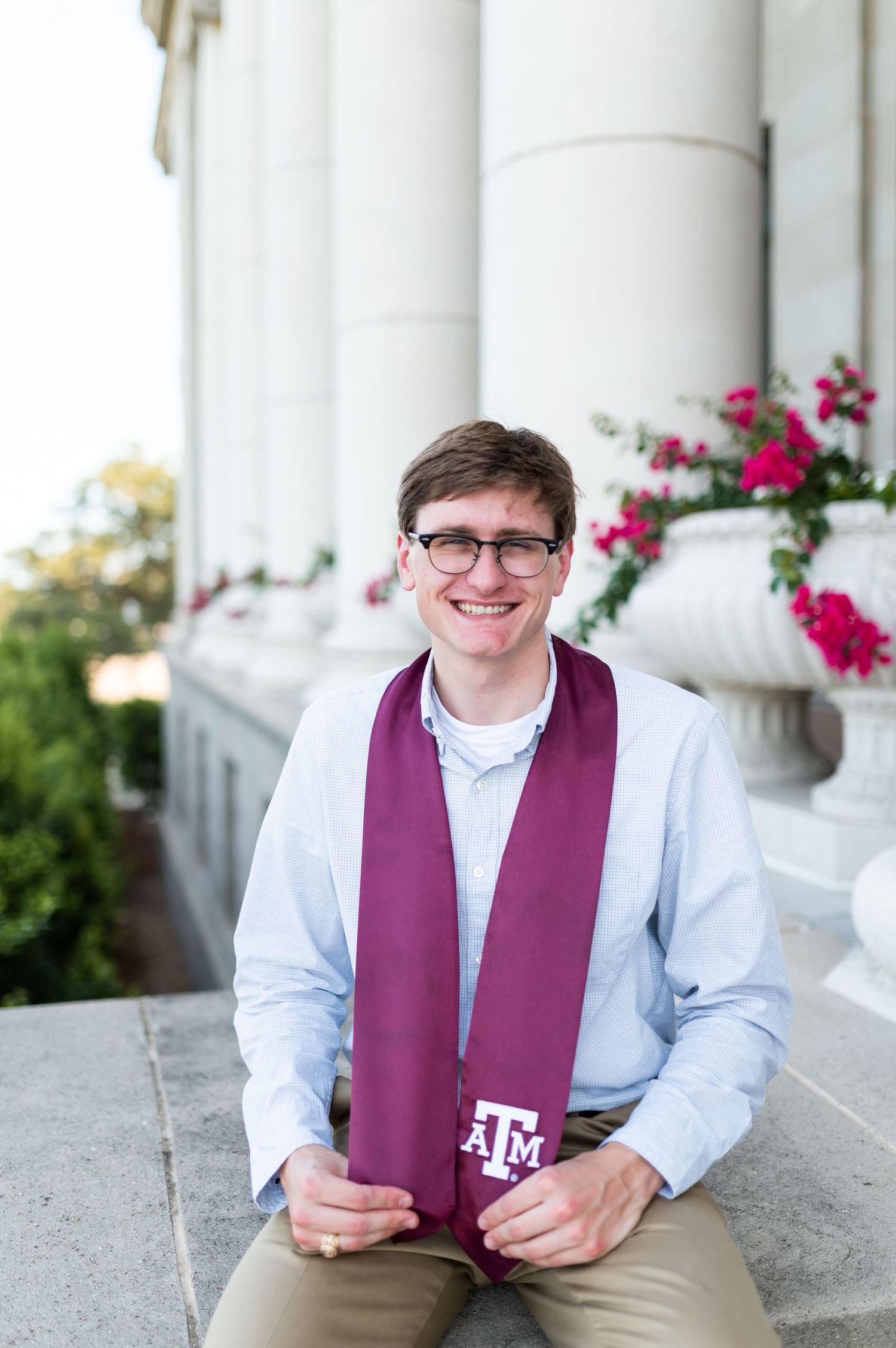 Smiling male Texas A&M graduate holds maroon ATM stole while sitting on steps near floral columns. Grad Photos at the Admin building