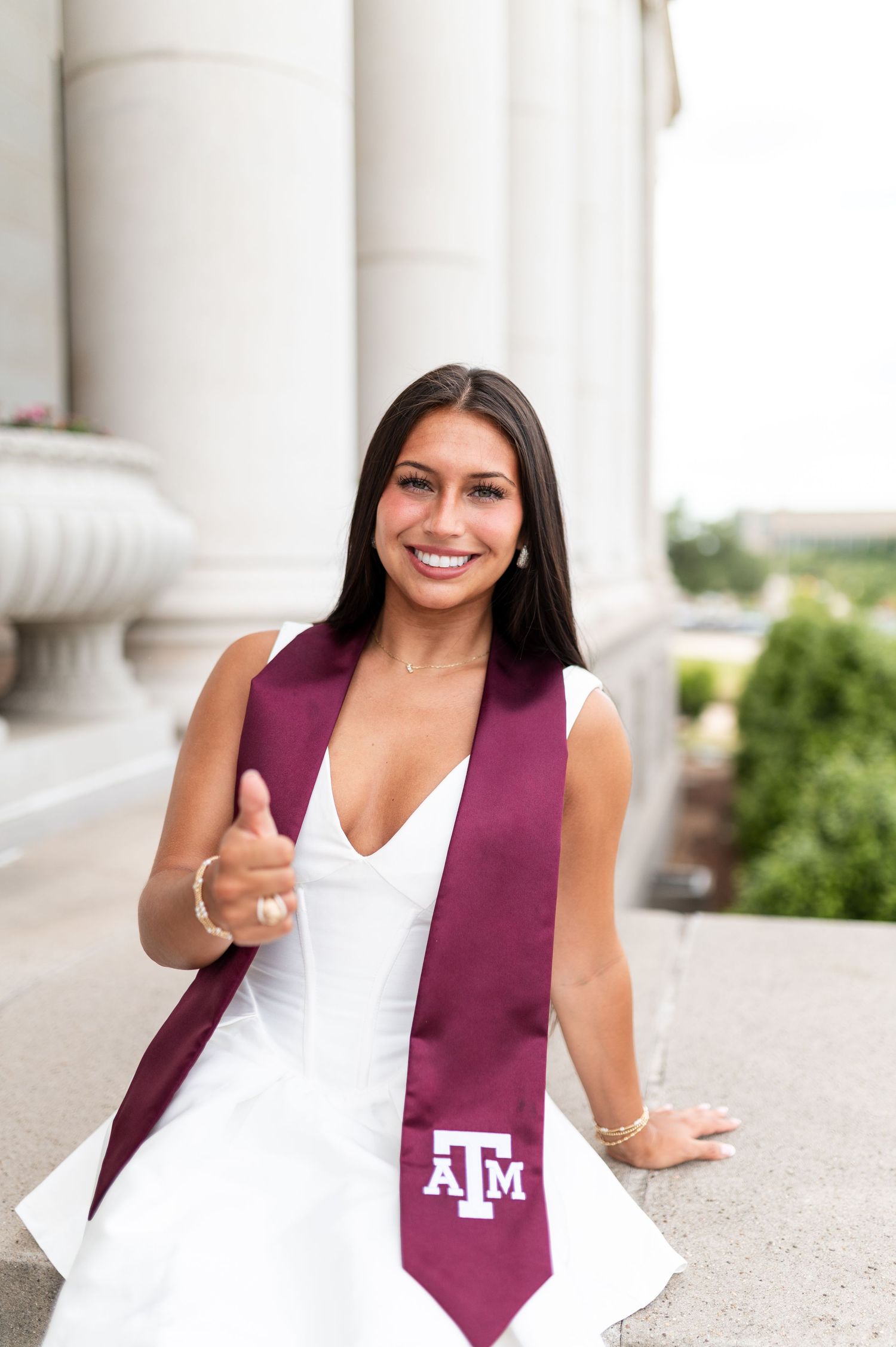 Texas A&M graduate in white dress with maroon stole giving thumbs up. Grad Photos at the Admin building