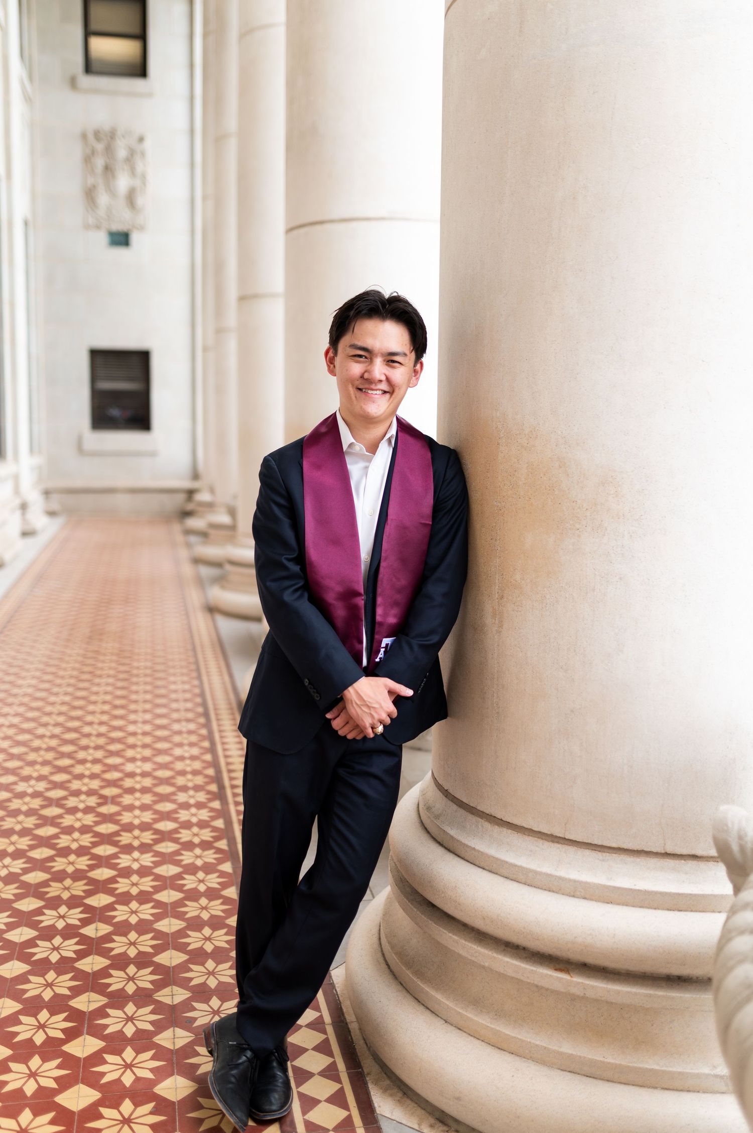 Young man in navy suit with maroon graduation stole smiles while leaning against campus column. Grad Photos at the Admin building