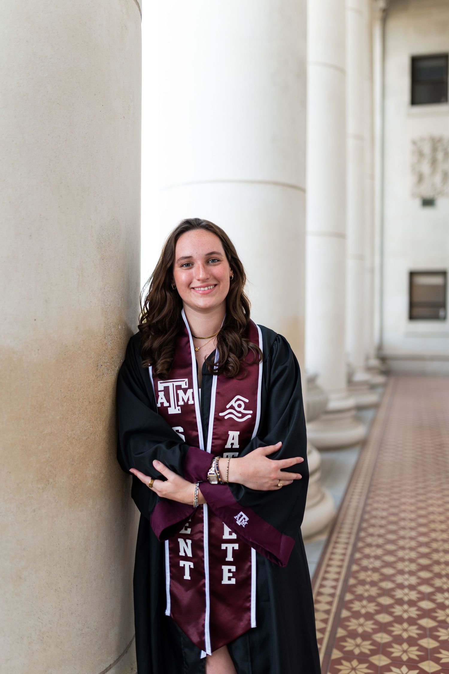 Smiling Texas A&M student athlete in graduation gown leans against column in campus corridor. Grad Photos at the Admin building