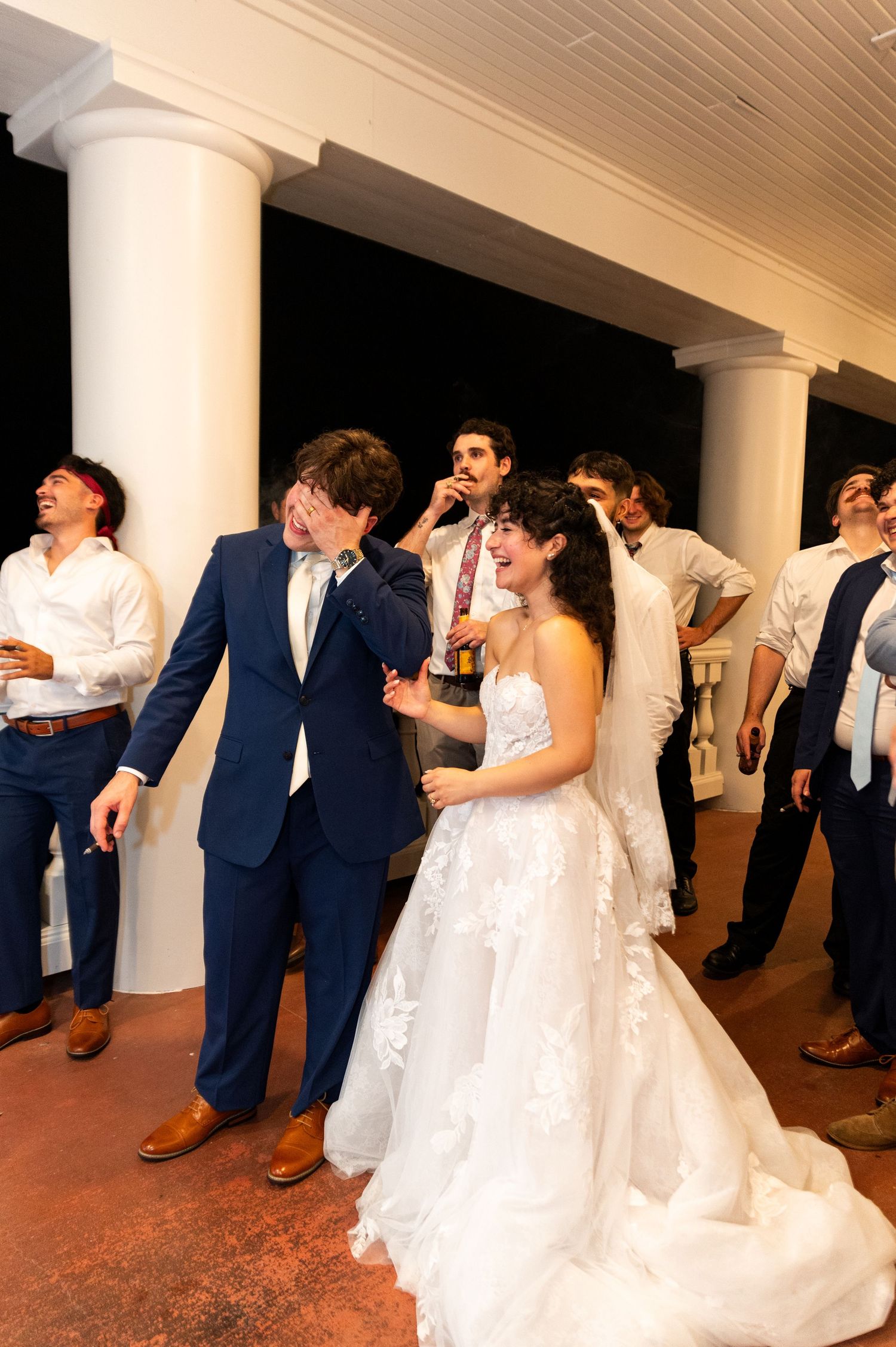 Bride and groom laughing with groomsmen in navy suits on venue porch during wedding celebration.