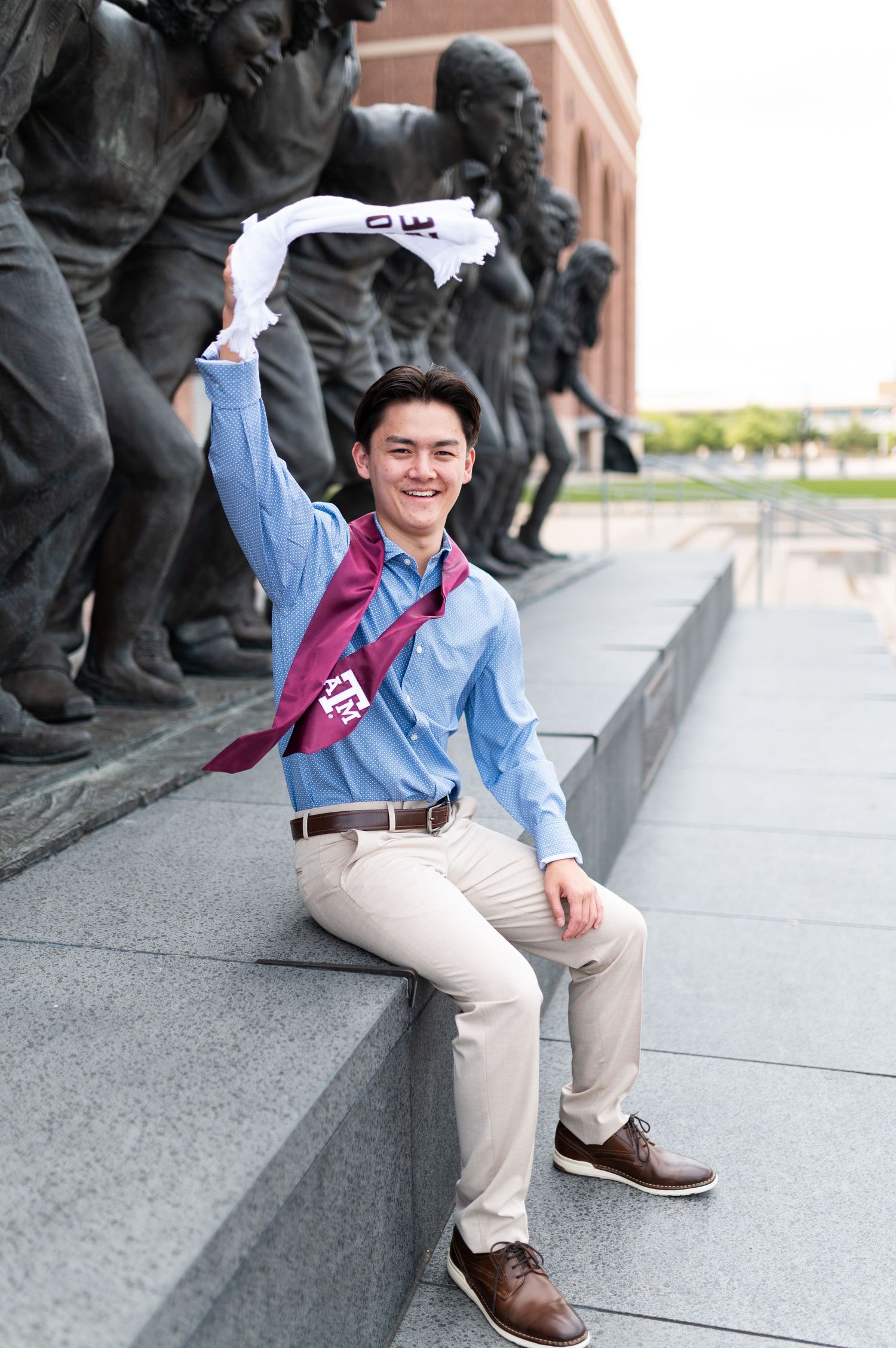 Excited young man waving towel wearing Texas A&M stole seated near campus bronze statues.