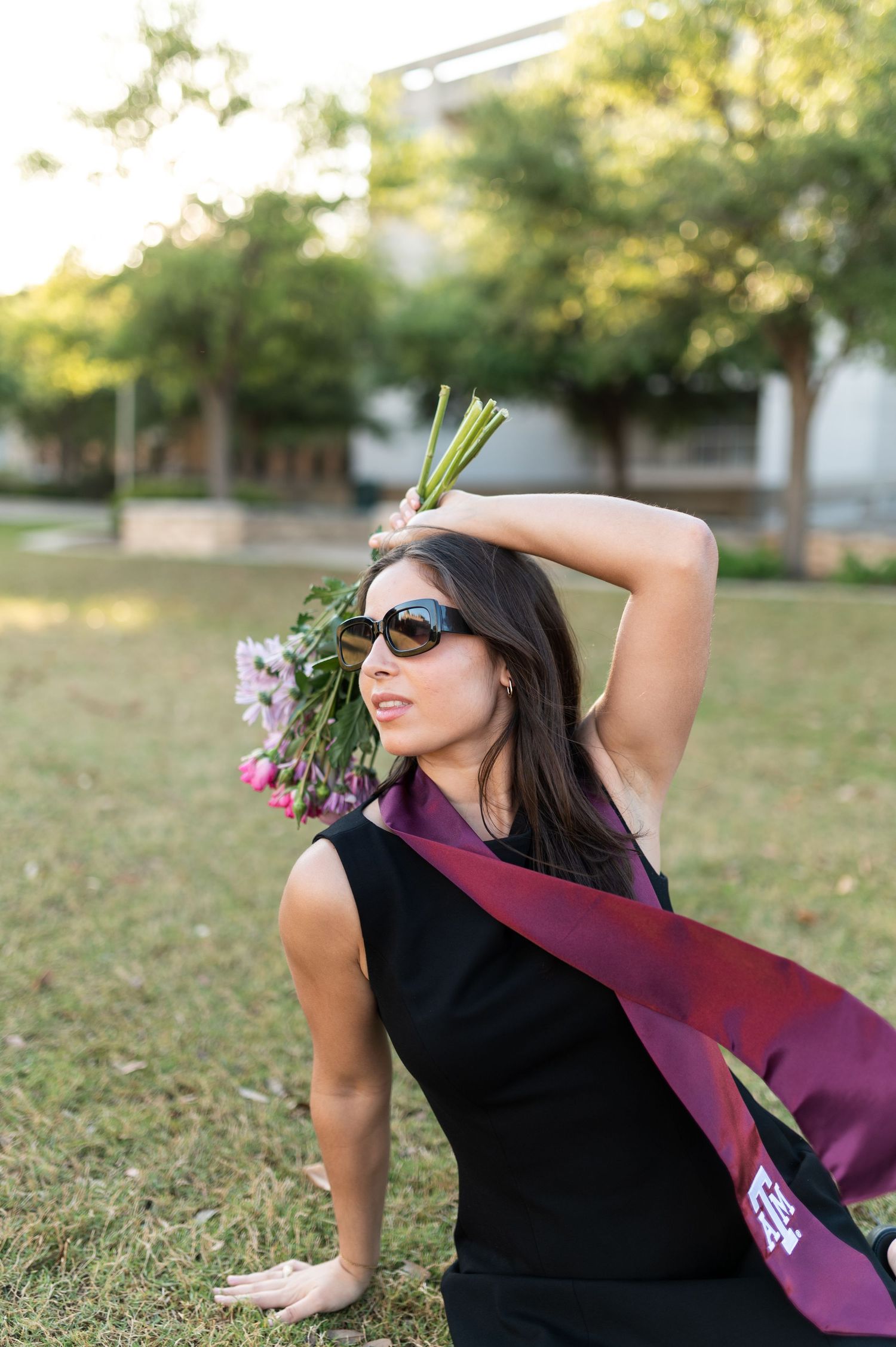 Texas A&M graduate sits on grass wearing maroon stole and sunglasses holding bouquet of pink flowers outdoors.