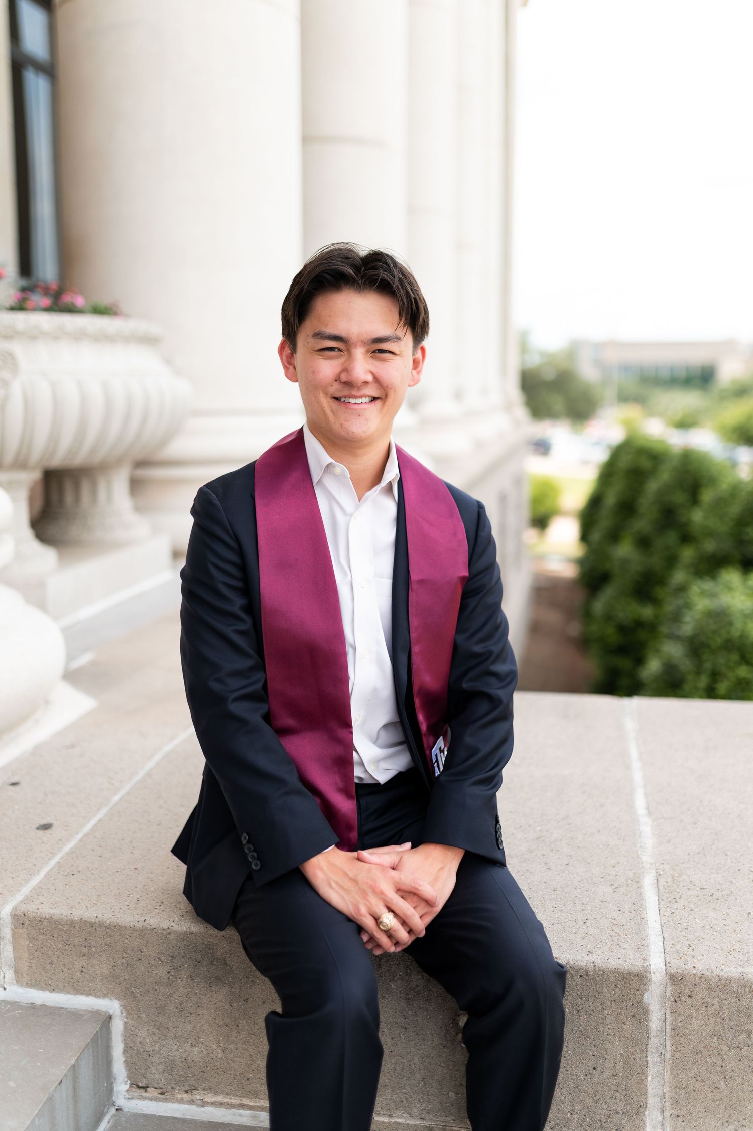 Smiling young man in navy suit with maroon graduation stole seated on stone steps at Texas A&M University. Grad Photos at the Admin building