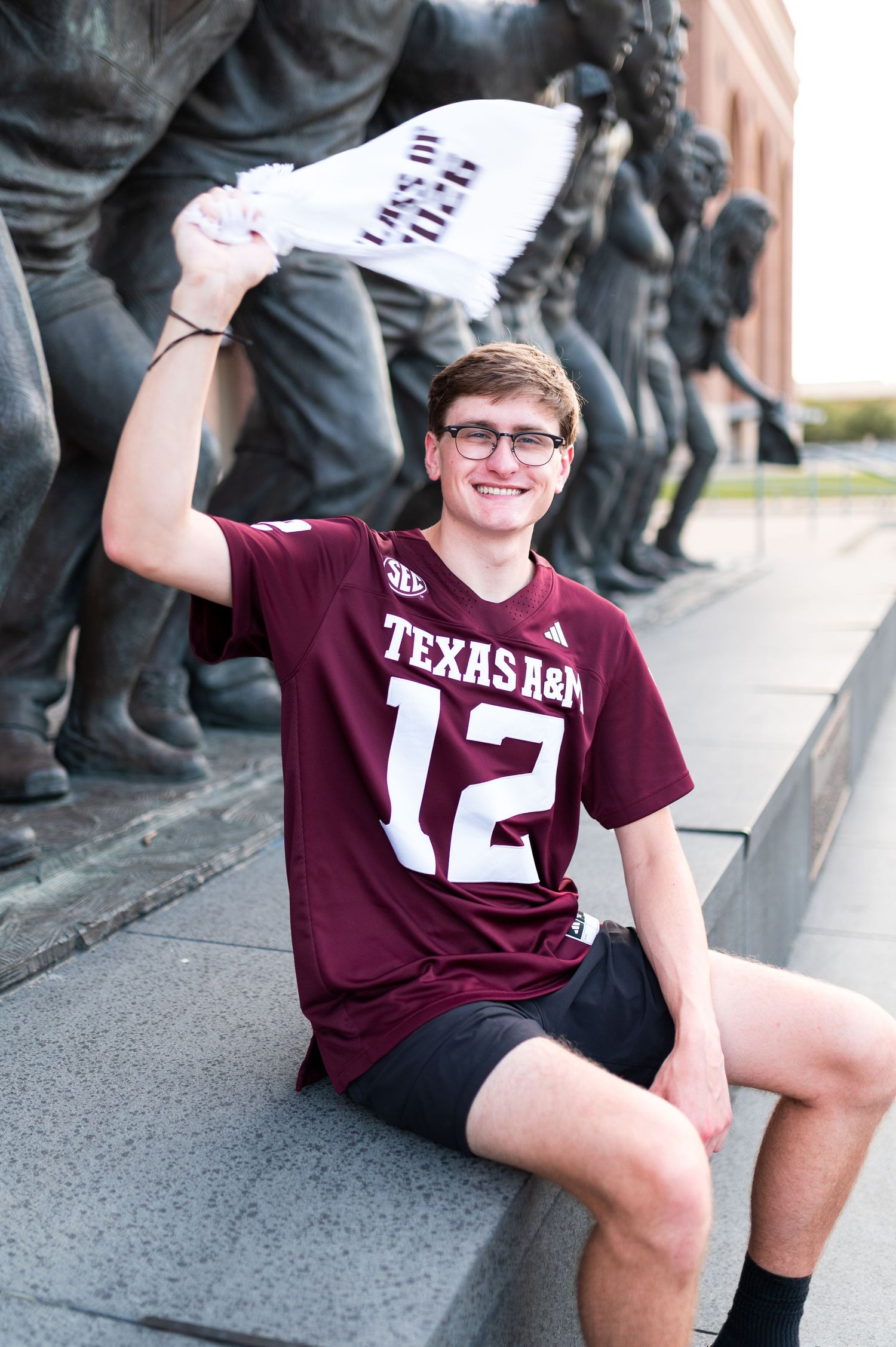 Young man in Texas A&M jersey waves towel beside bronze statues outside Kyle Field.