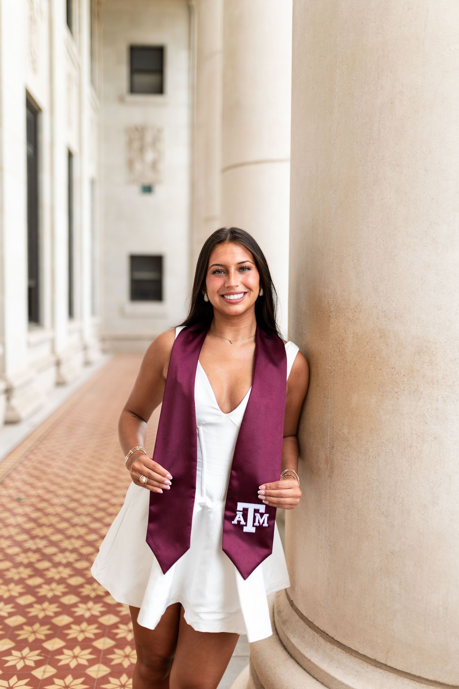 Smiling female Texas A&M grad in white dress holds maroon ATM stole against campus column. Grad Photos at the Texas A&M Admin building