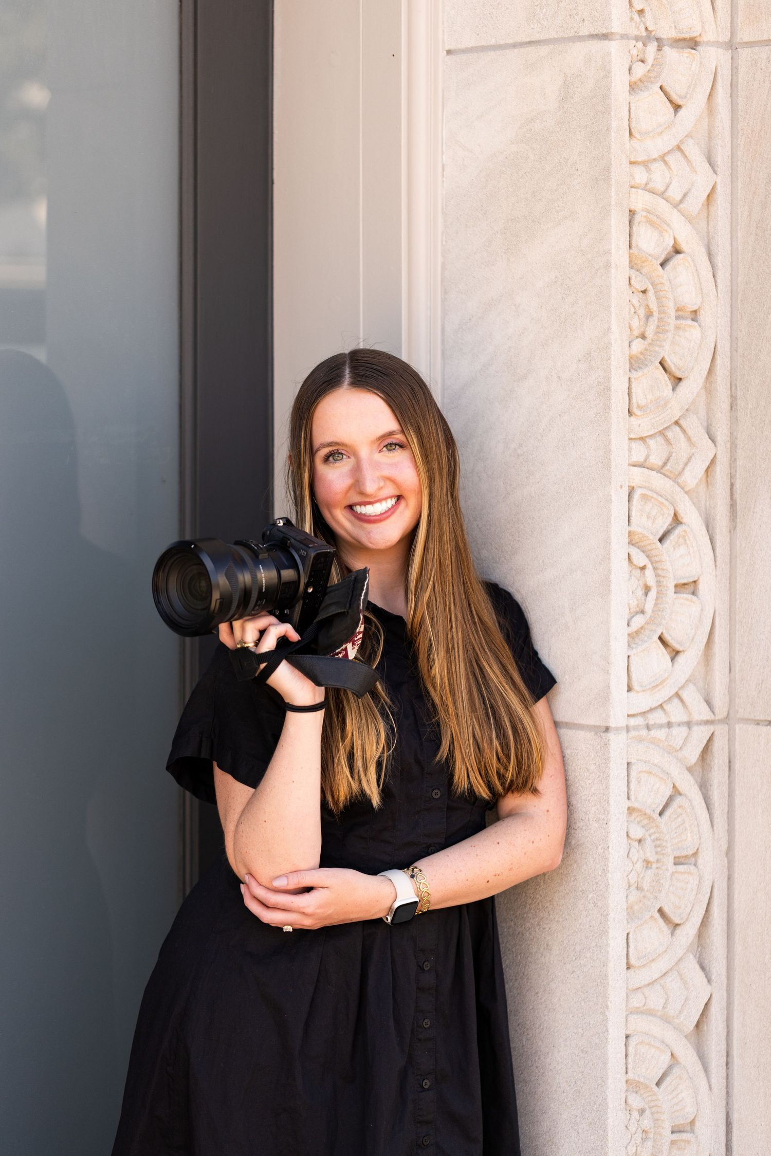 Smiling woman with long brown hair in a black dress holds a professional camera while leaning against an ornate carved stone wall.
