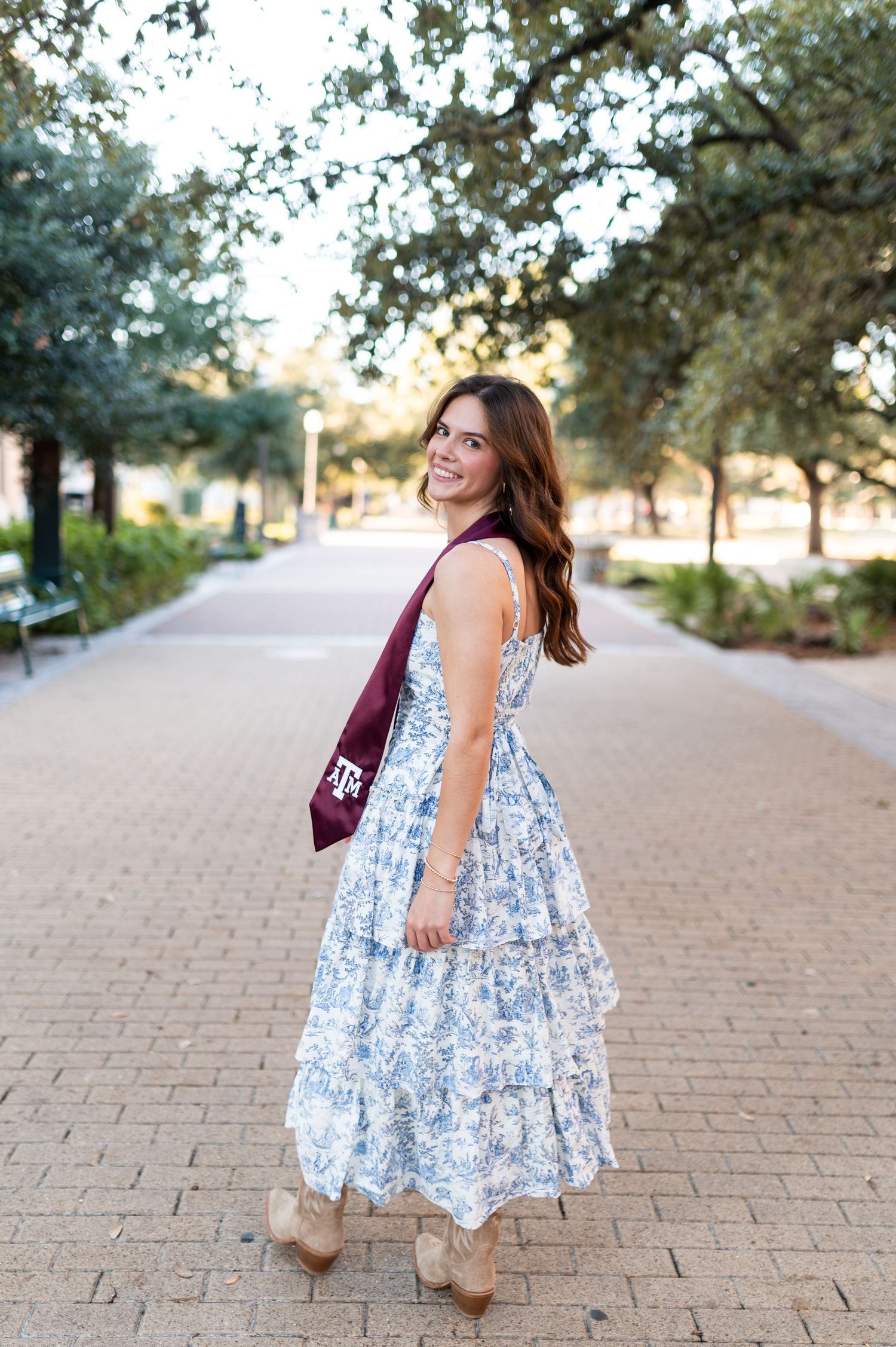 Texas A&M graduate twirls in blue floral dress with maroon graduation stole on brick campus walkway.