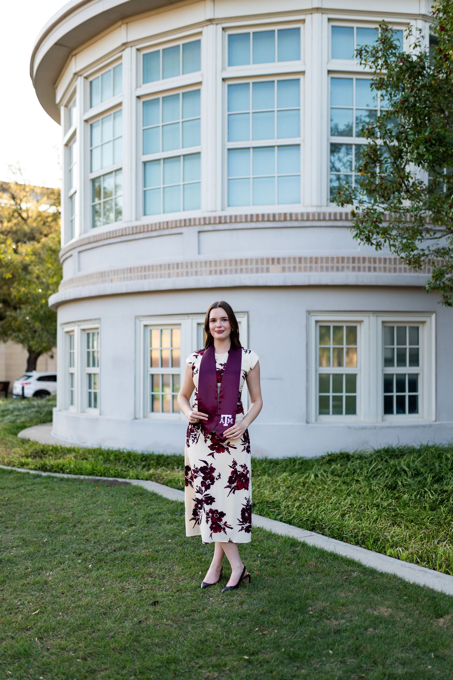 Smiling woman in floral midi dress holds Texas A&M items while standing on grass near round building.