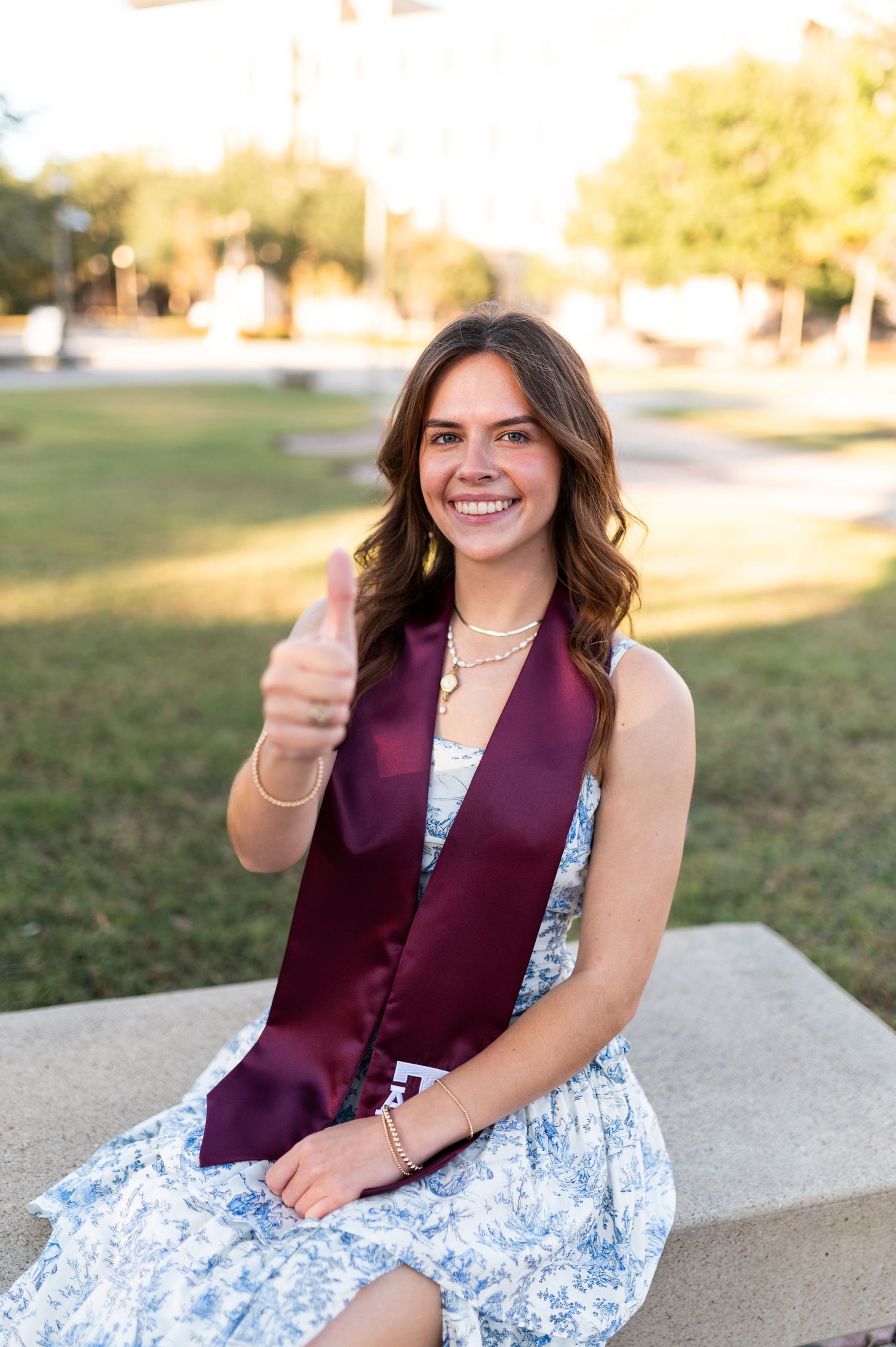 Happy Texas A&M graduate in blue floral dress and maroon stole gives thumbs up seated on stone bench.