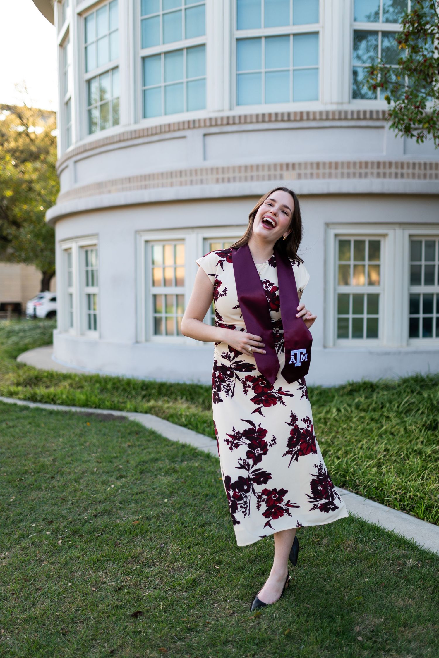Laughing Texas A&M graduate in floral dress holds maroon graduation stole outside curved building.