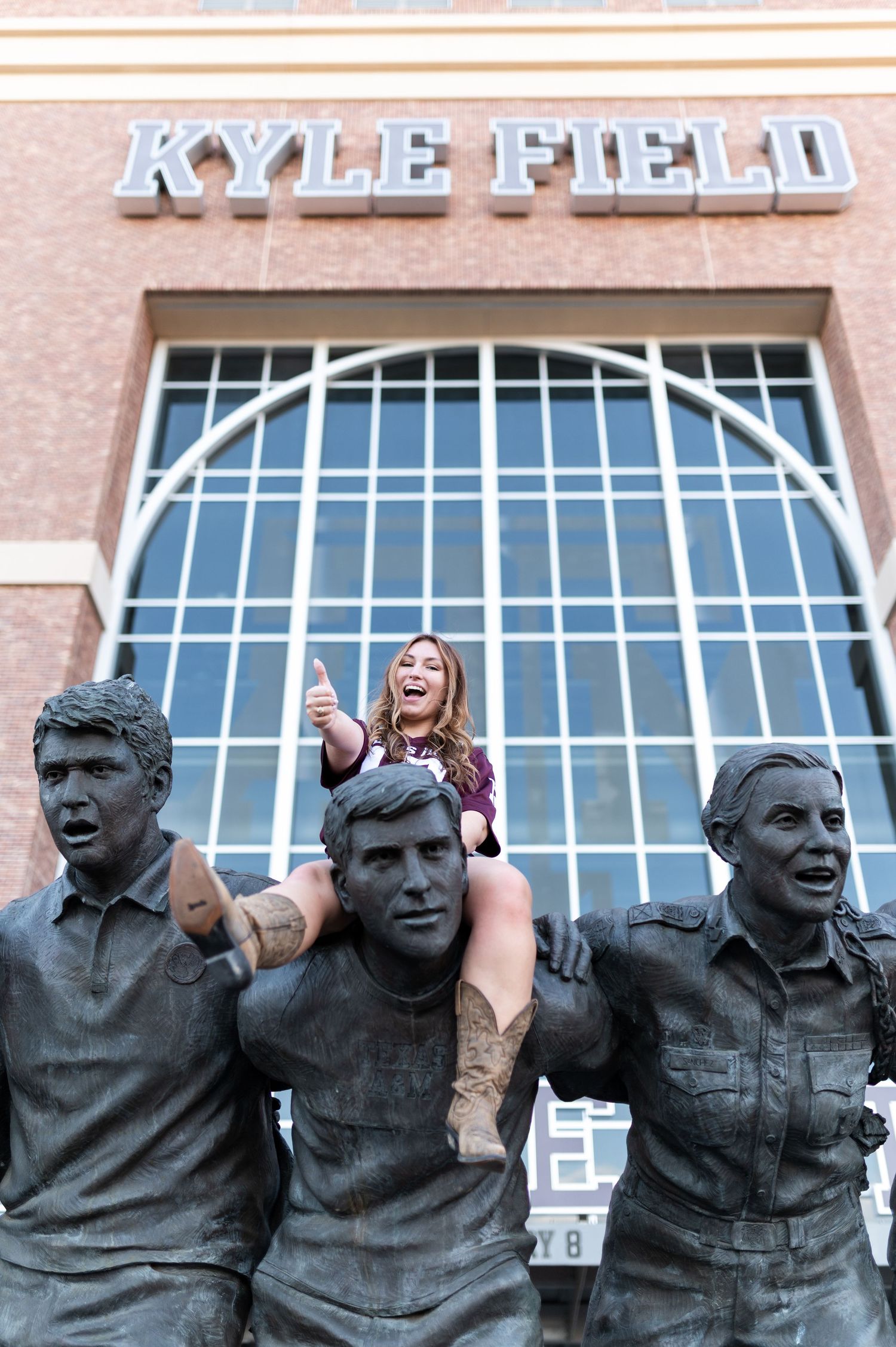Woman sits on statue shoulders at Kyle Field, Texas A&M, giving thumbs up.