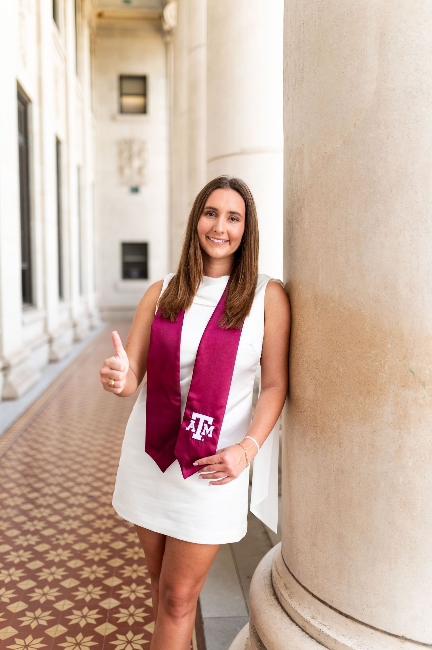 Texas A&M female graduate in white dress gives thumbs up leaning against column wearing maroon ATM sash. Grad Photos at the Texas A&M Admin