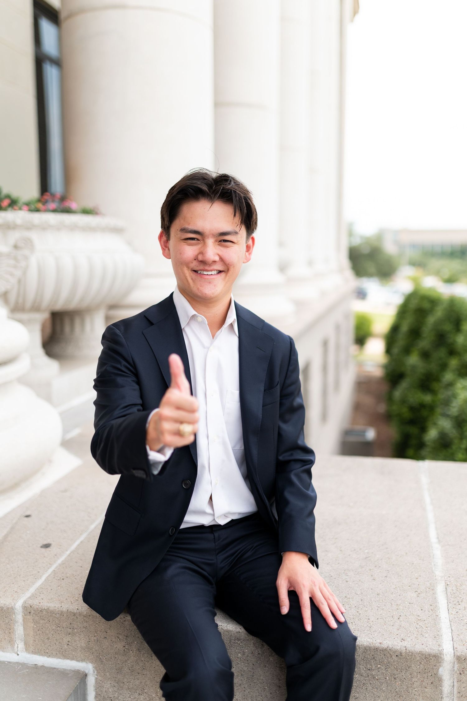 Cheerful young man in navy blazer giving thumbs up while seated on stone ledge at Texas A&M University. Grad Photos at the Admin building
