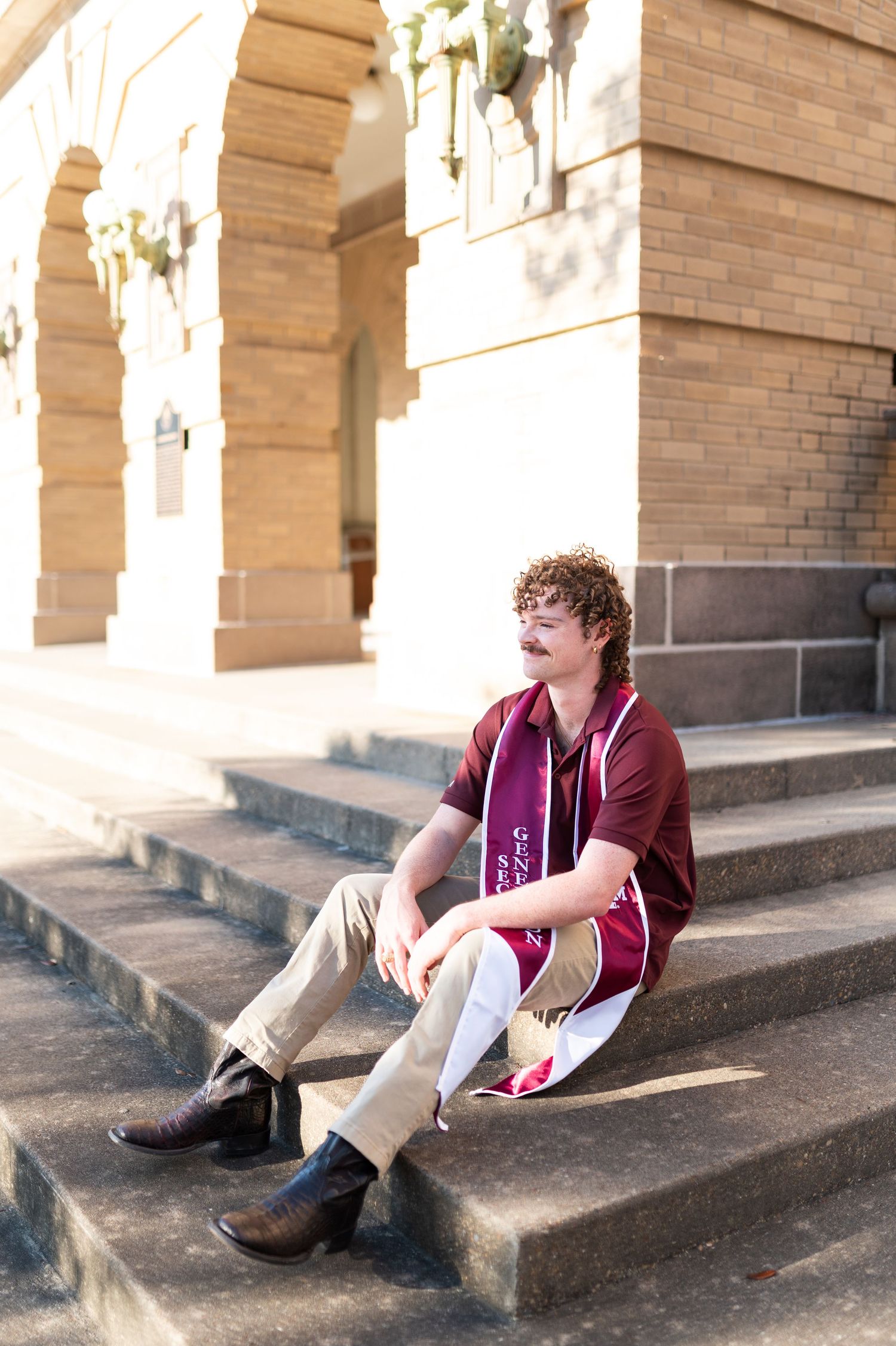 Young man with curly hair and graduation stole sits relaxed on stone steps.