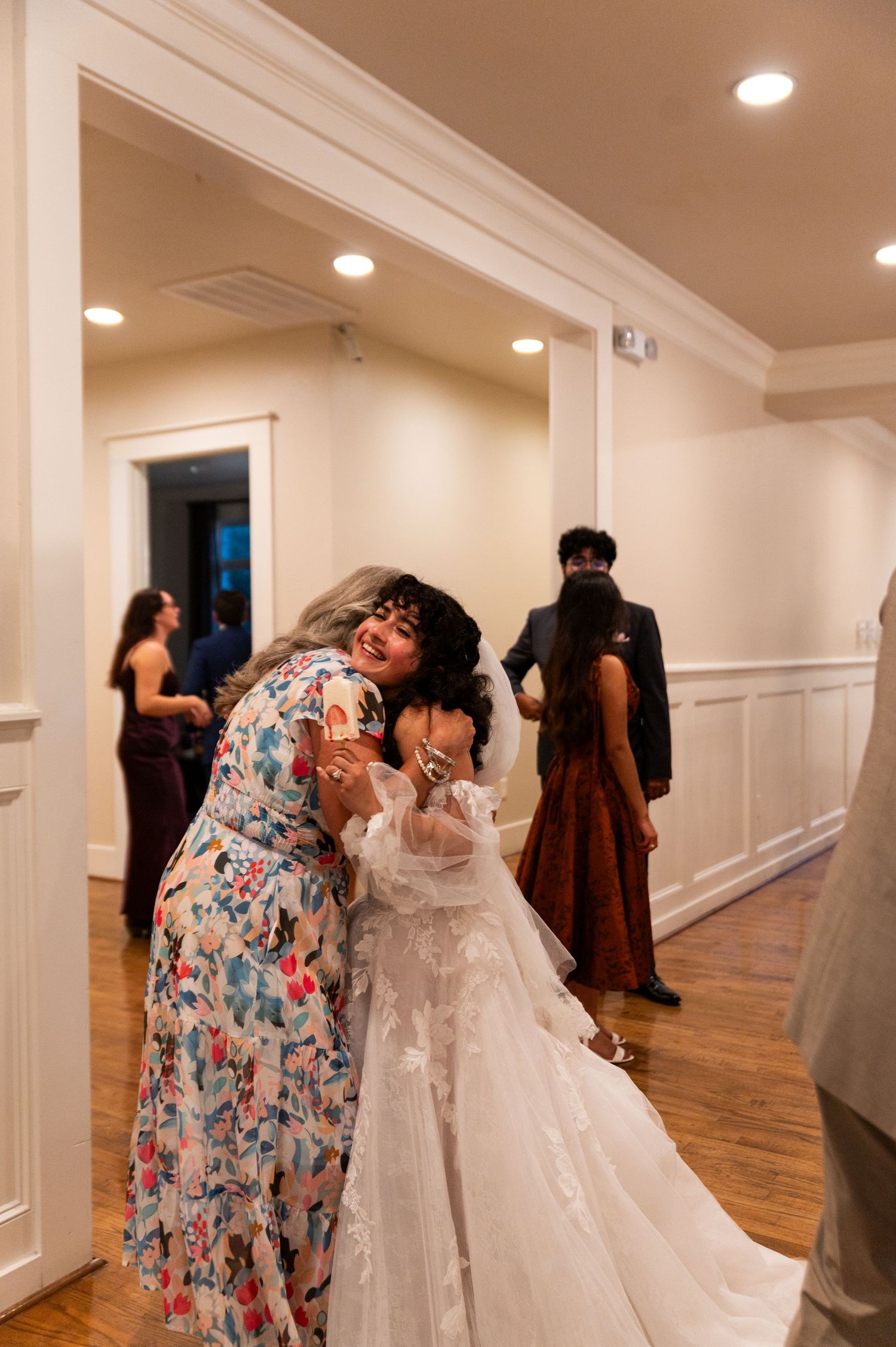 Bride in lace gown receives warm hug from guest in floral dress at wedding reception.